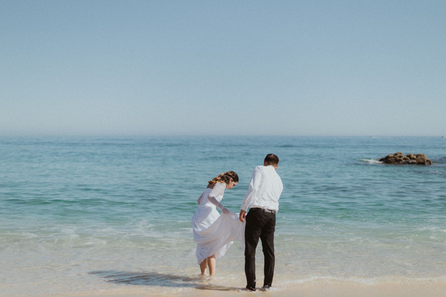 A romantic beach photo of a couple in formal attire walking along turquoise waters and sandy shores.