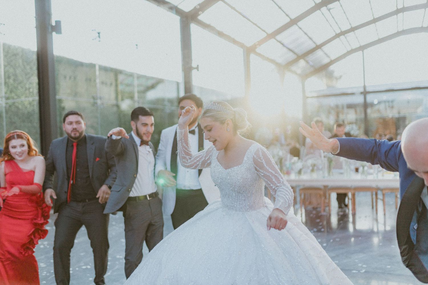 People celebrate at a joyful wedding reception in a covered venue as sunlight streams through the windows.