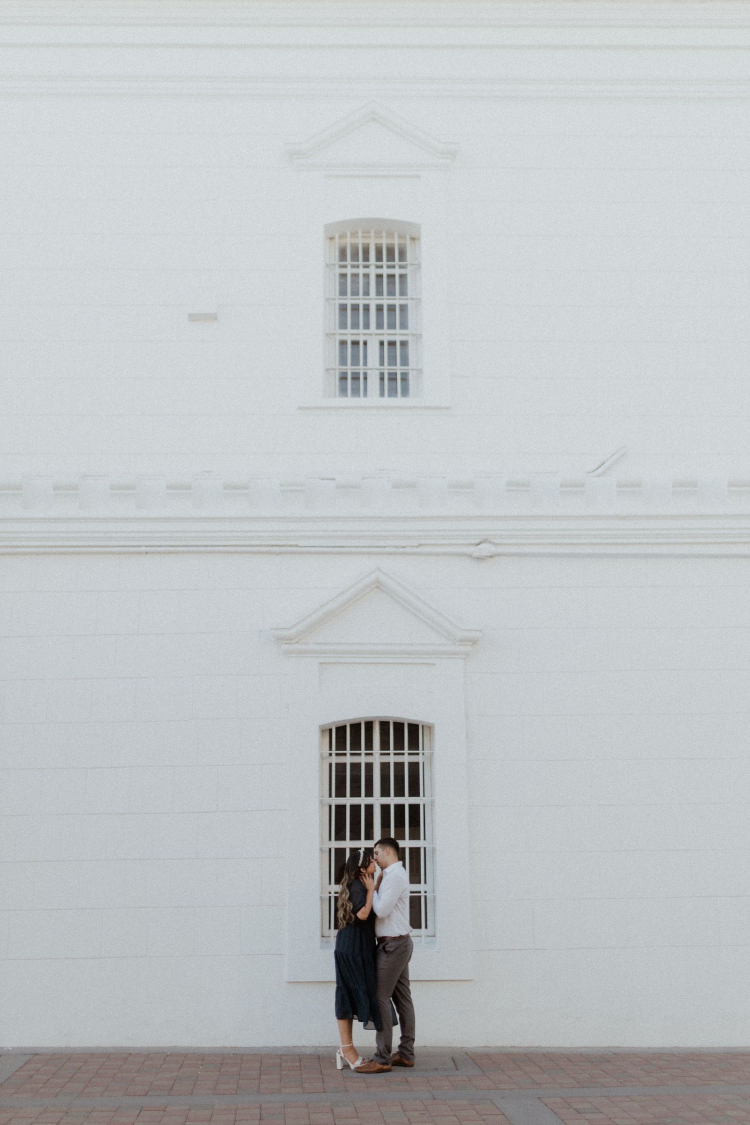 A couple shares an intimate moment against a white colonial building with barred windows.