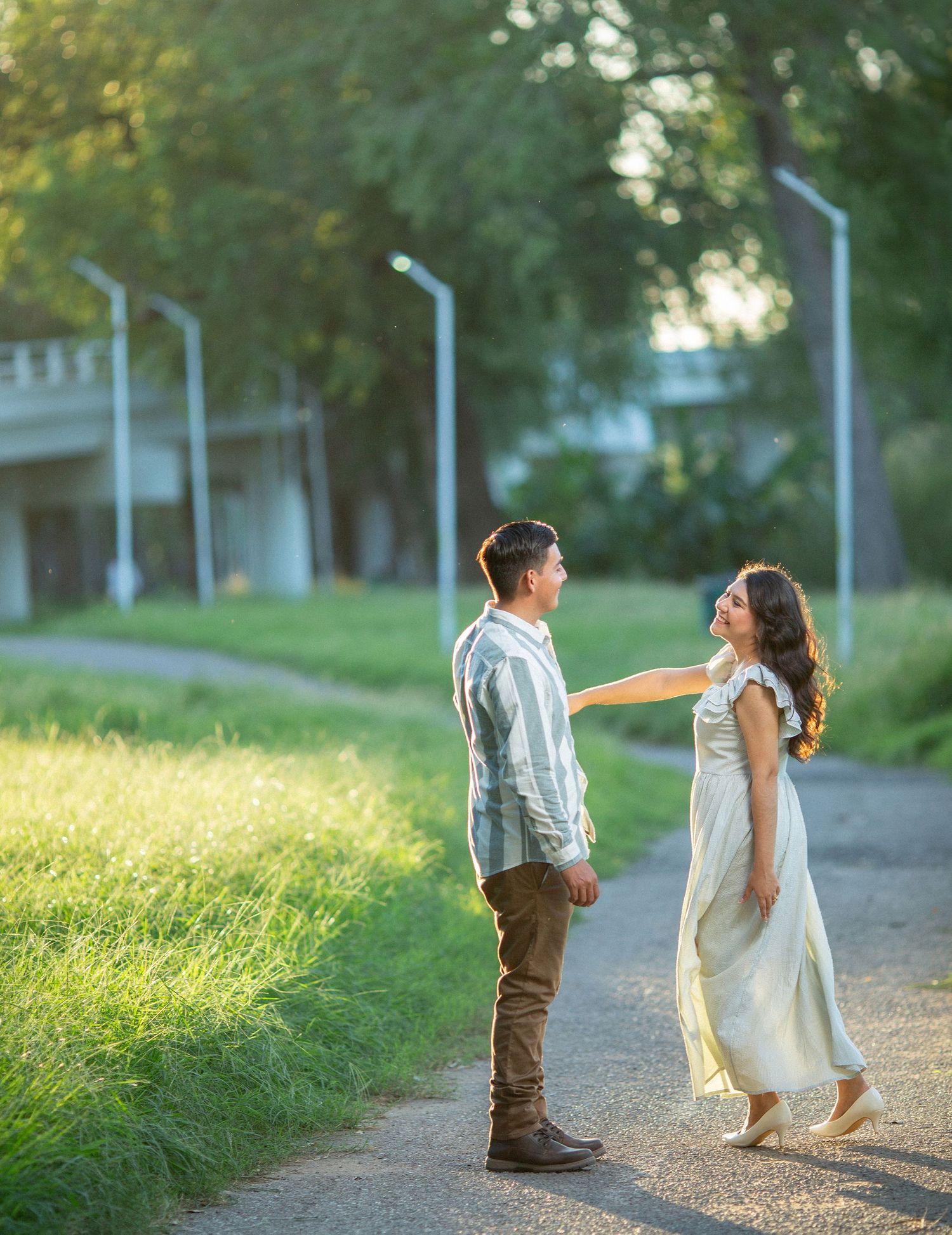 A couple shares a tender moment at sunset on a tree-lined path with glowing streetlamps in the background.