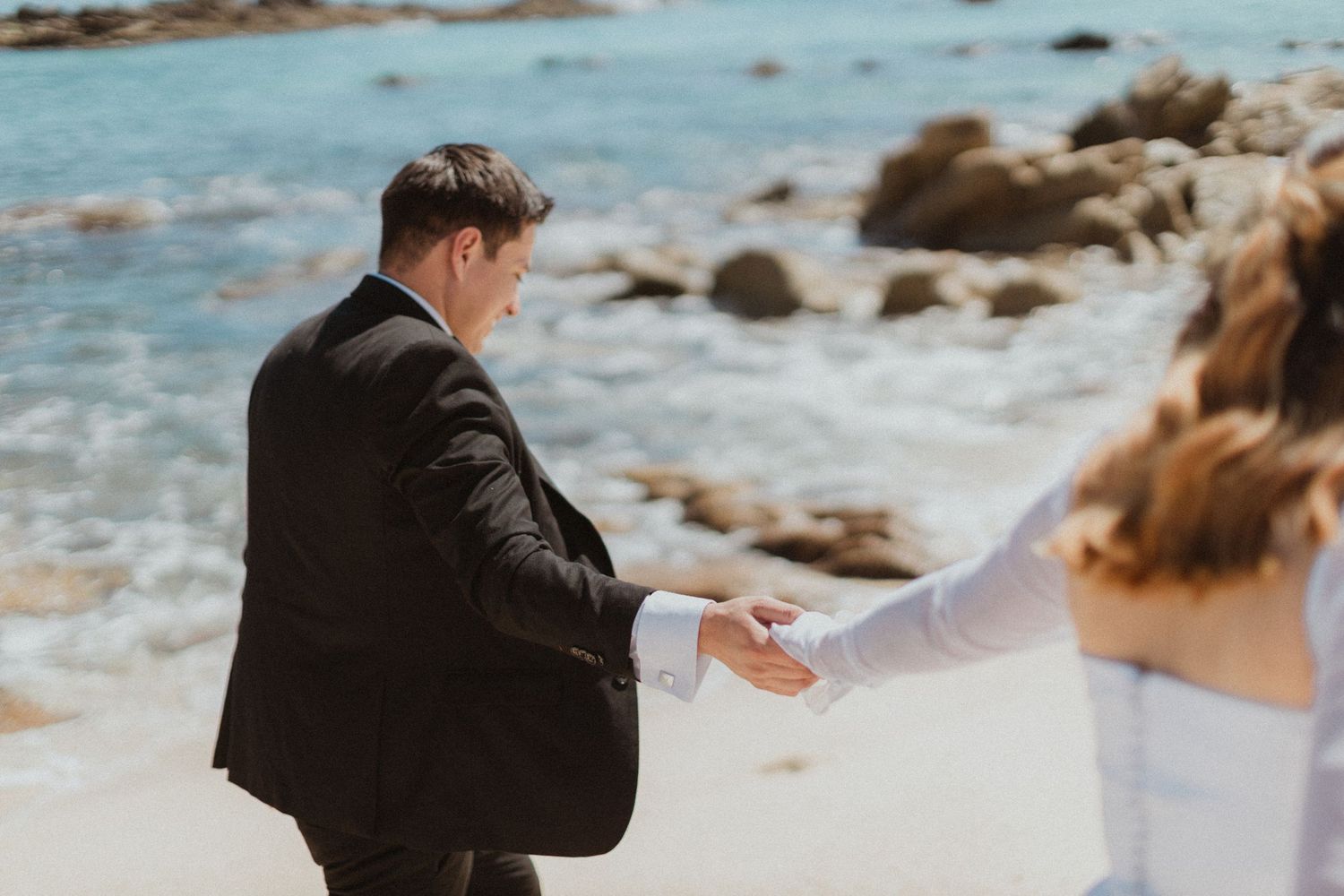 Couple in wedding attire holds hands on beach rocks.