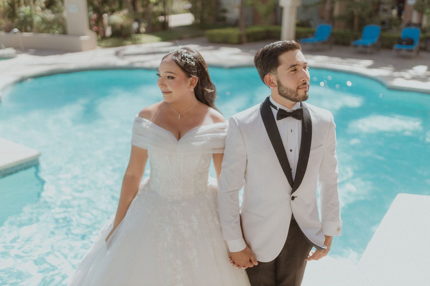 A bride and groom in formal attire stand hand-in-hand by a swimming pool.