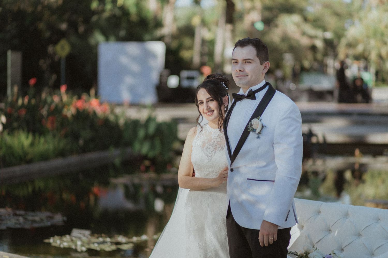 A couple poses in formal wedding attire by a reflective pond in a garden setting.