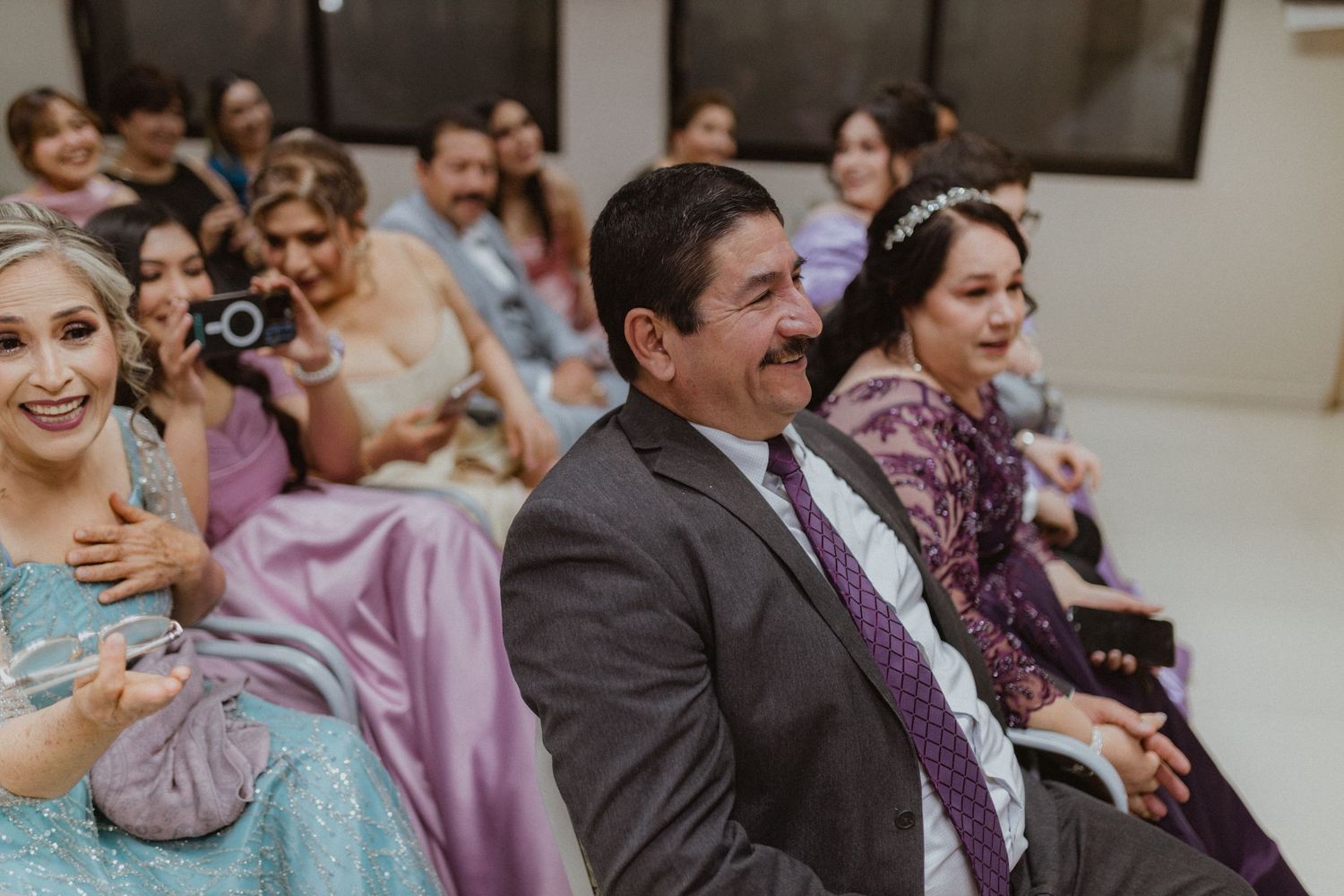Wedding guests laugh and smile during ceremony in decorated room with purple accents.