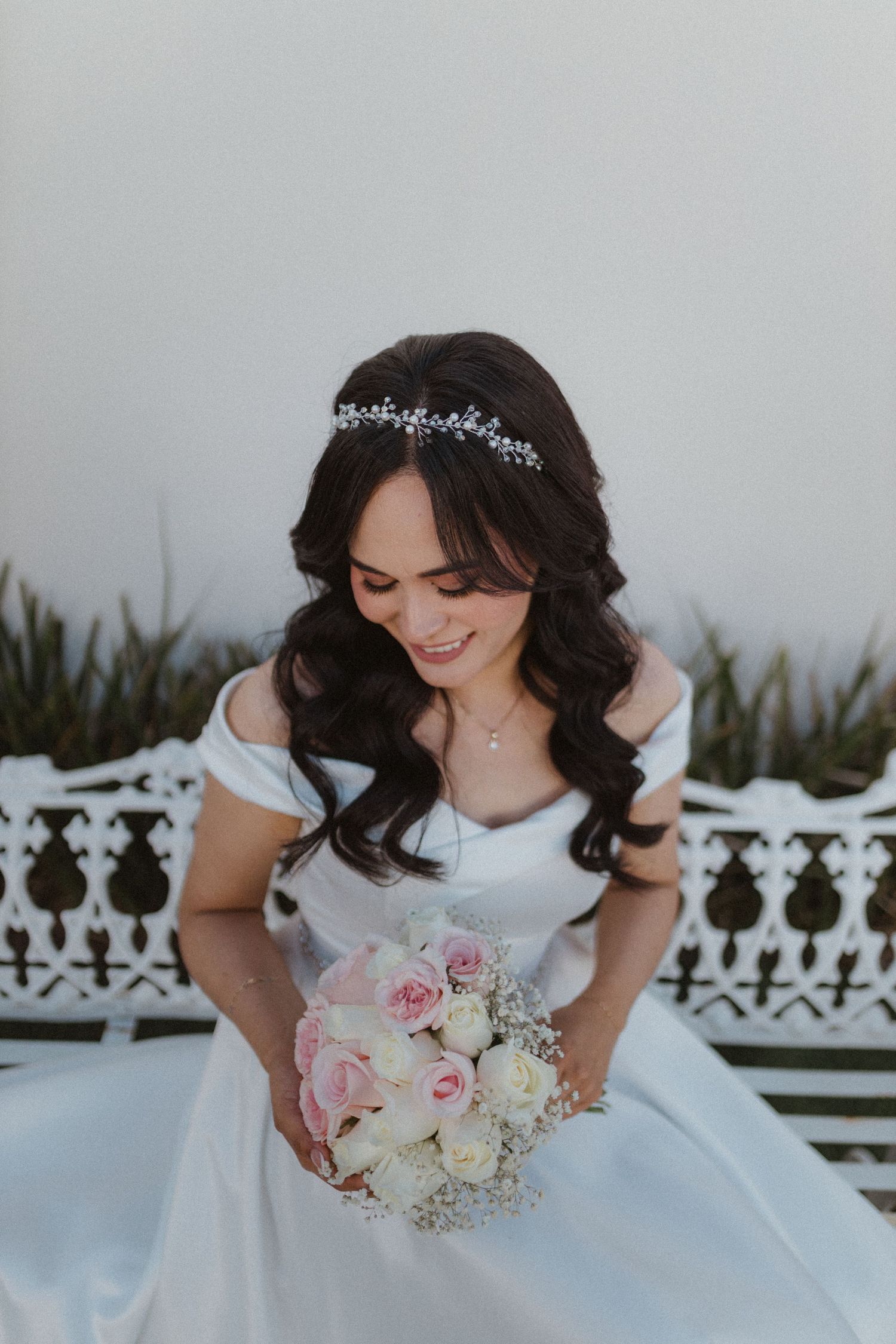 A close-up shot of a bride in a white dress holding a pink and white bouquet on a decorative bench.