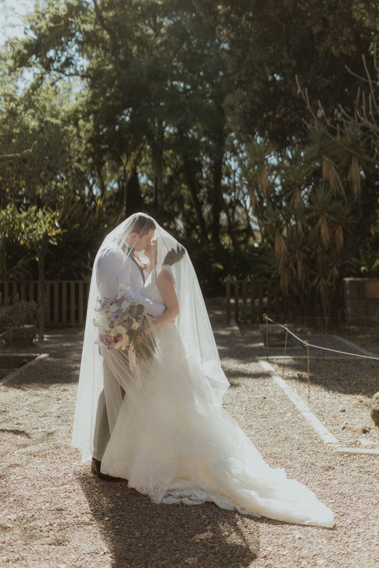 Bride in flowing white gown and veil holds purple bouquet in sunlit garden setting.