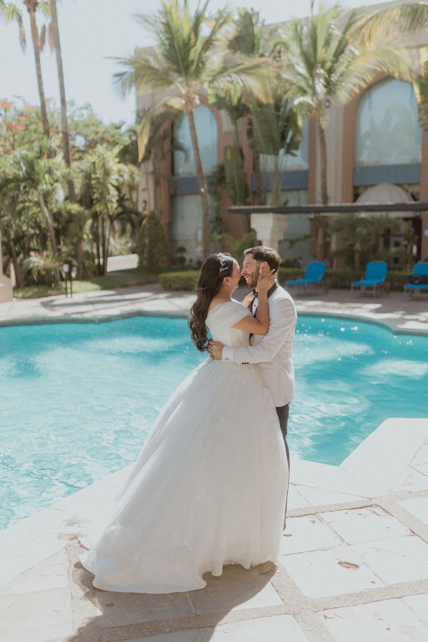 Elegant wedding couple dancing by tropical pool with palm trees and arched architecture.