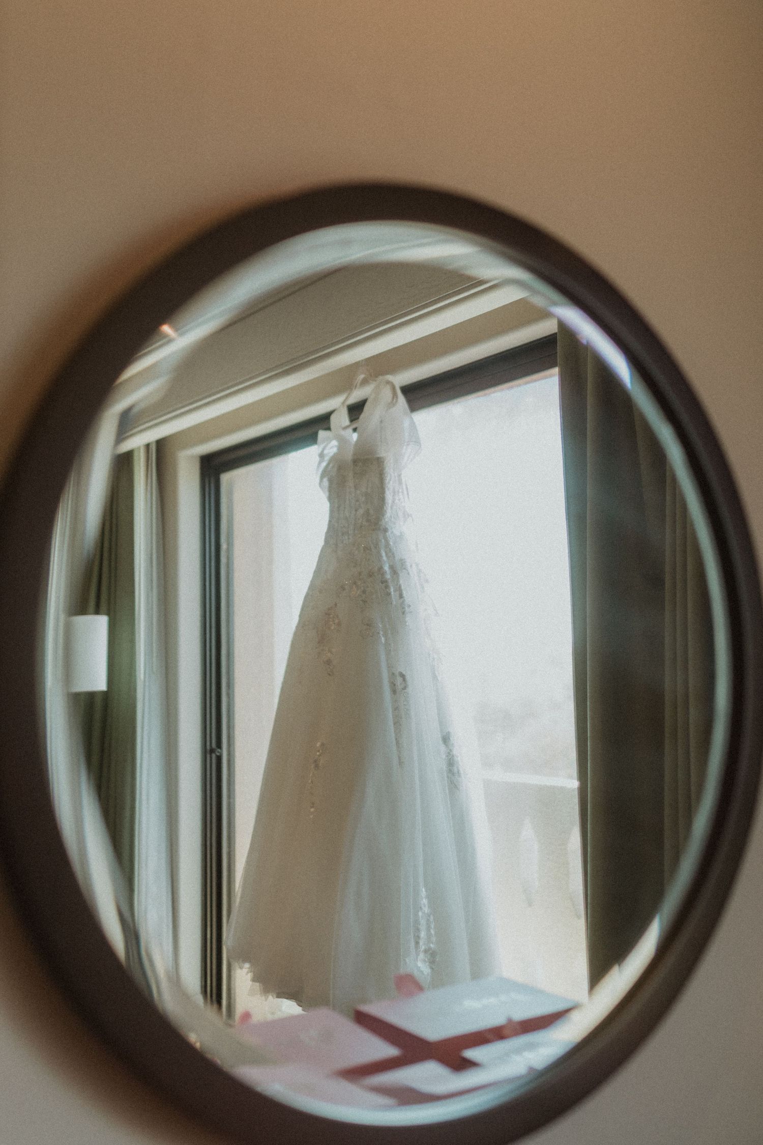 A wedding dress hangs in front of a window, reflected in an oval mirror.