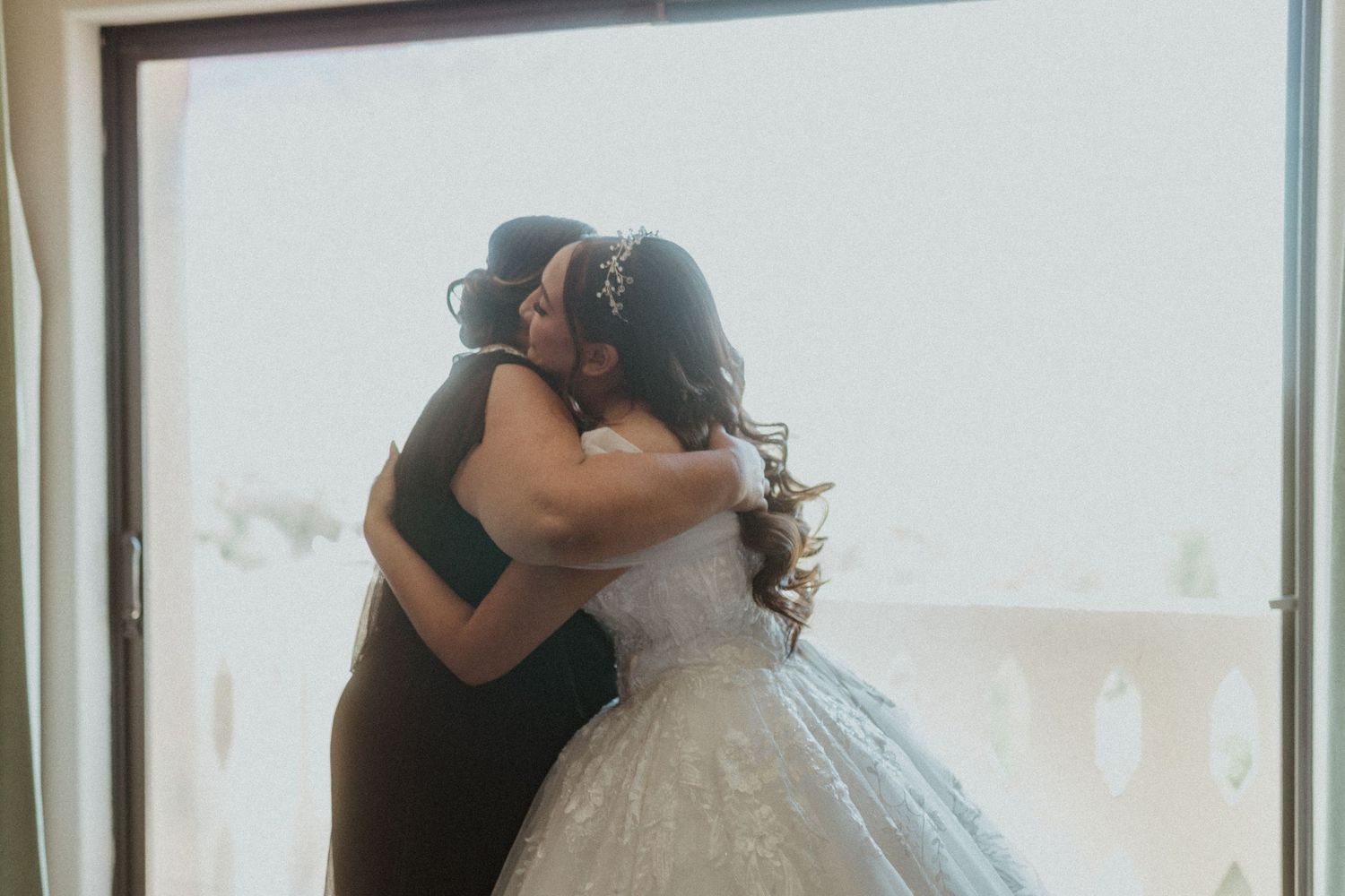 Two figures share an intimate embrace near a bright window during a wedding ceremony.