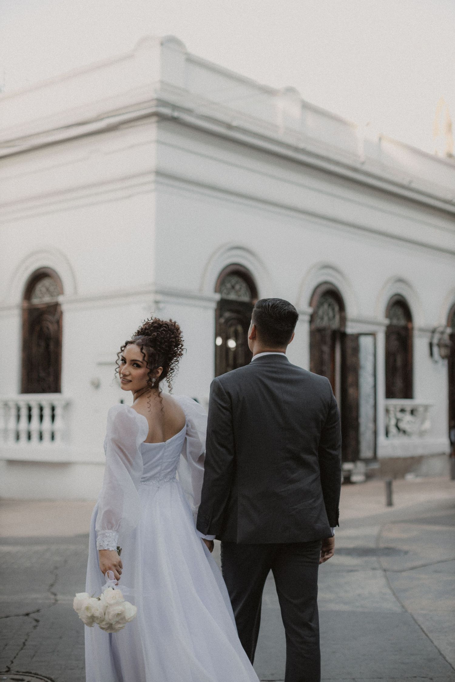 A couple in formal wedding attire standing outside a classic white colonial building with arched windows.
