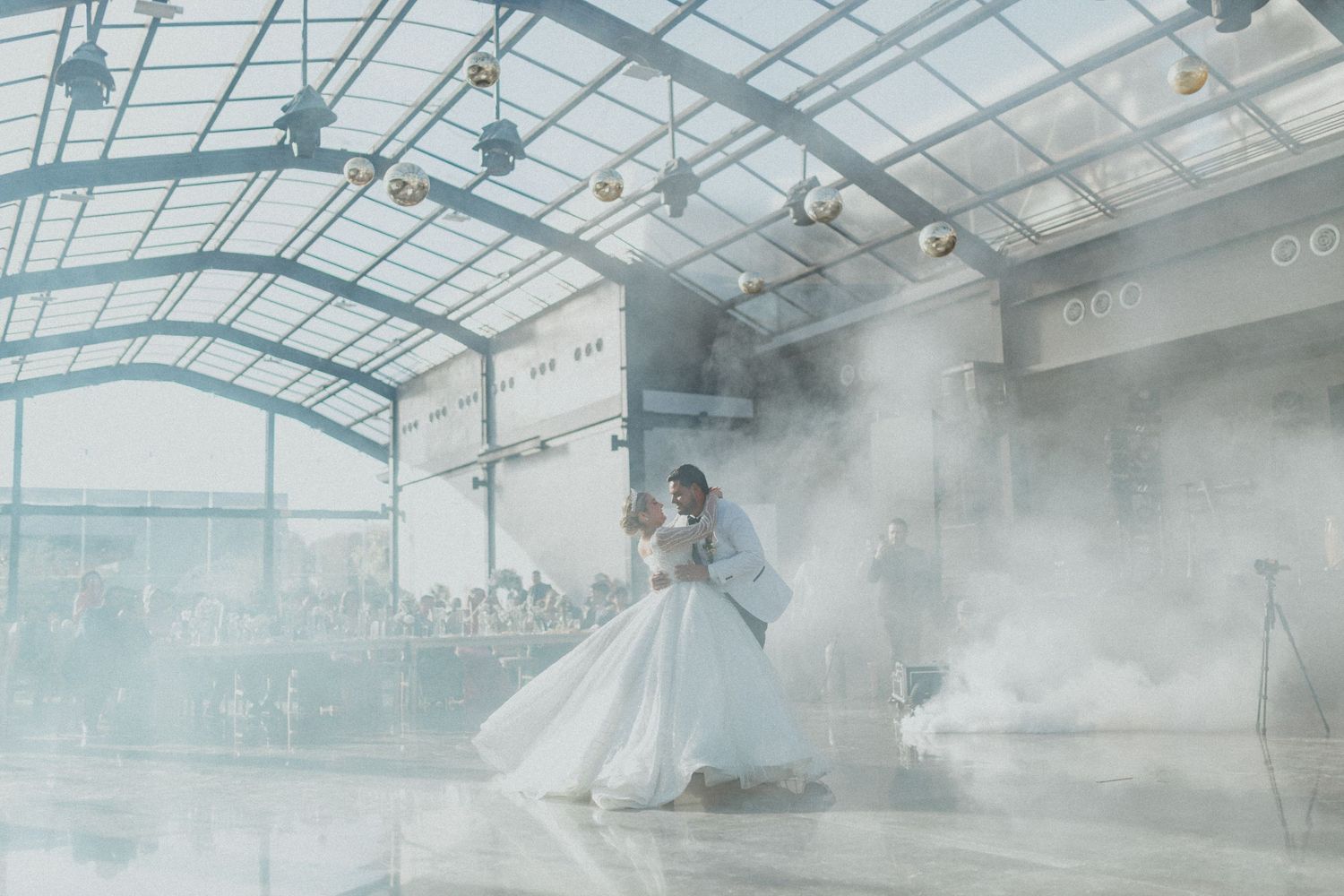 A misty greenhouse interior with glass ceiling and plants silhouetted against bright natural light.