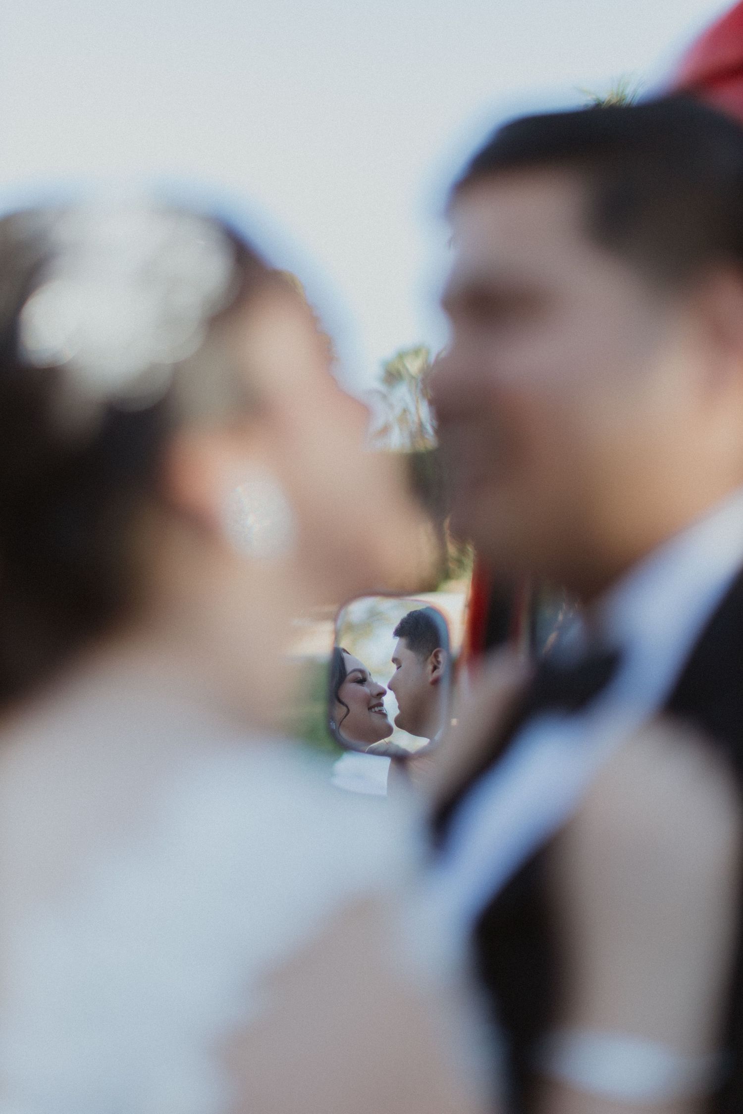 Artistic wedding photo showing couple's reflection in car mirror with bokeh effect.