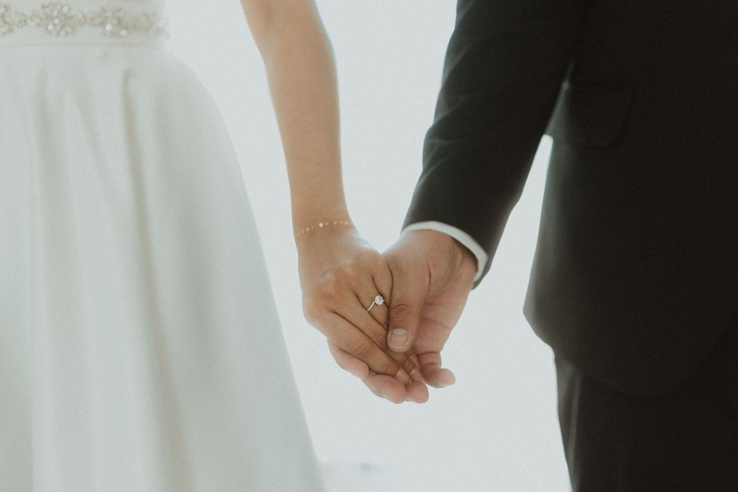Close-up of newlyweds holding hands, showing white dress and black suit details.