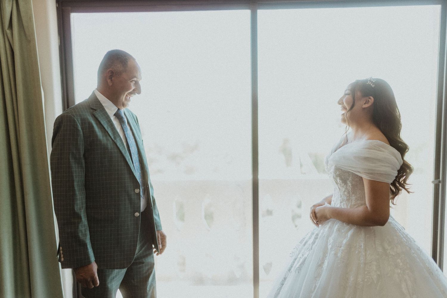 A wedding first look scene unfolds by large windows with natural light streaming in.