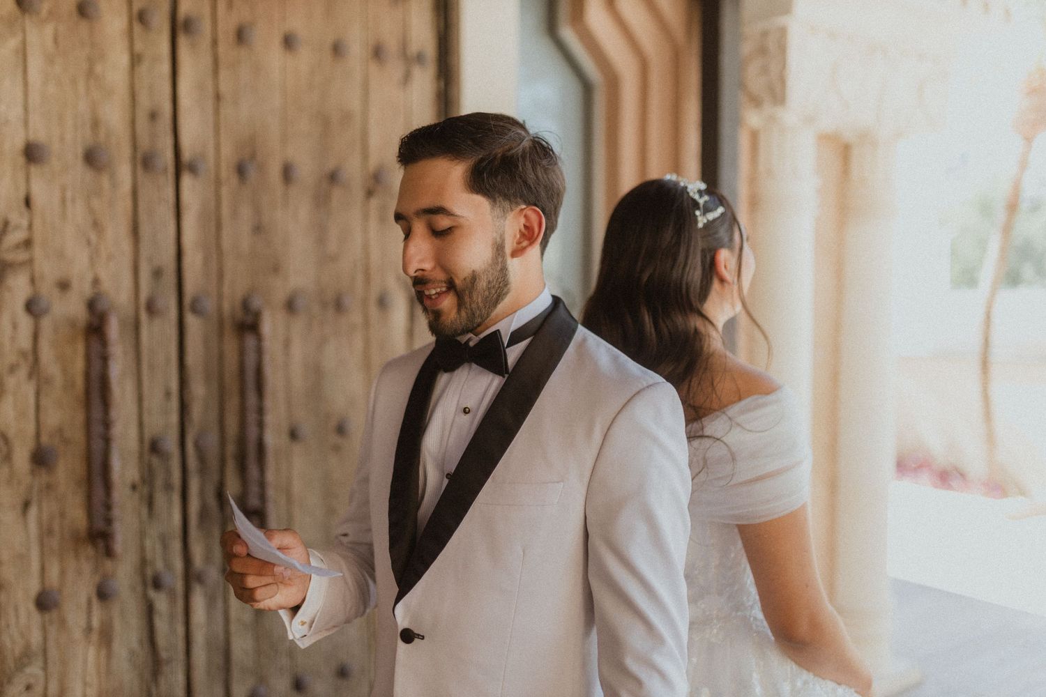 A groom in a white tuxedo reads a letter while his bride stands behind him during their first look.