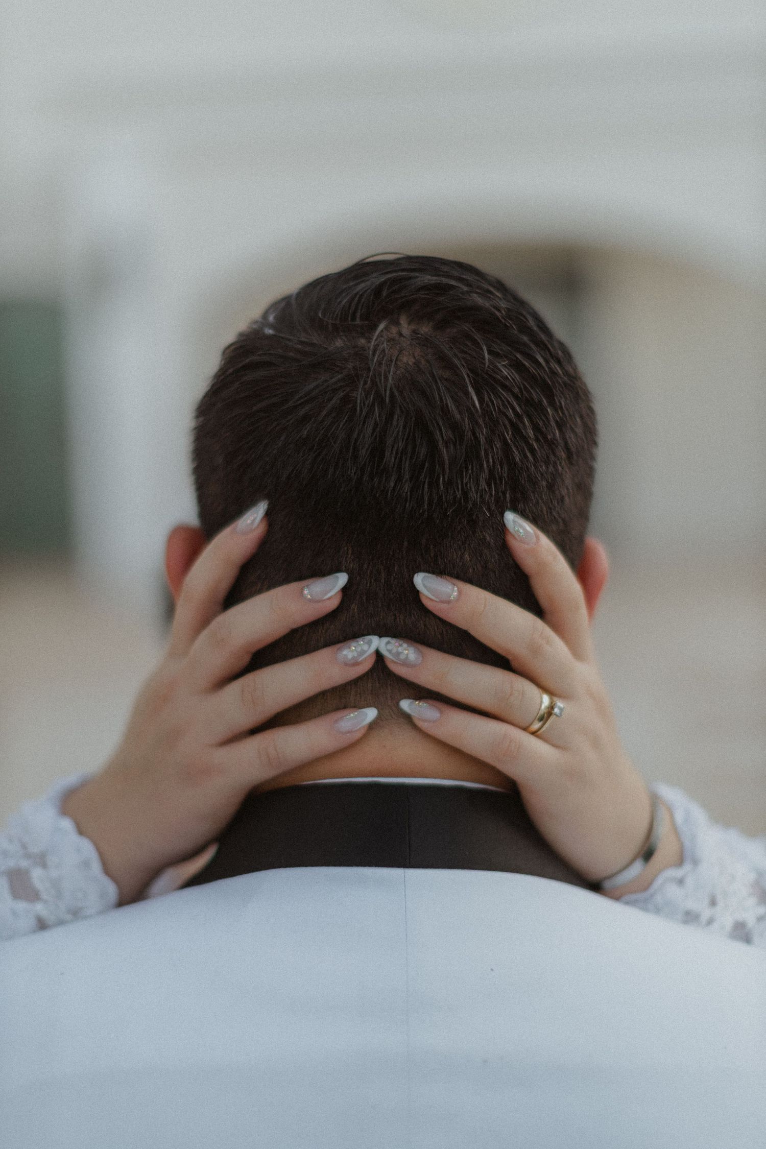 Detail shot of manicured hands with wedding ring holding groom's head from behind.