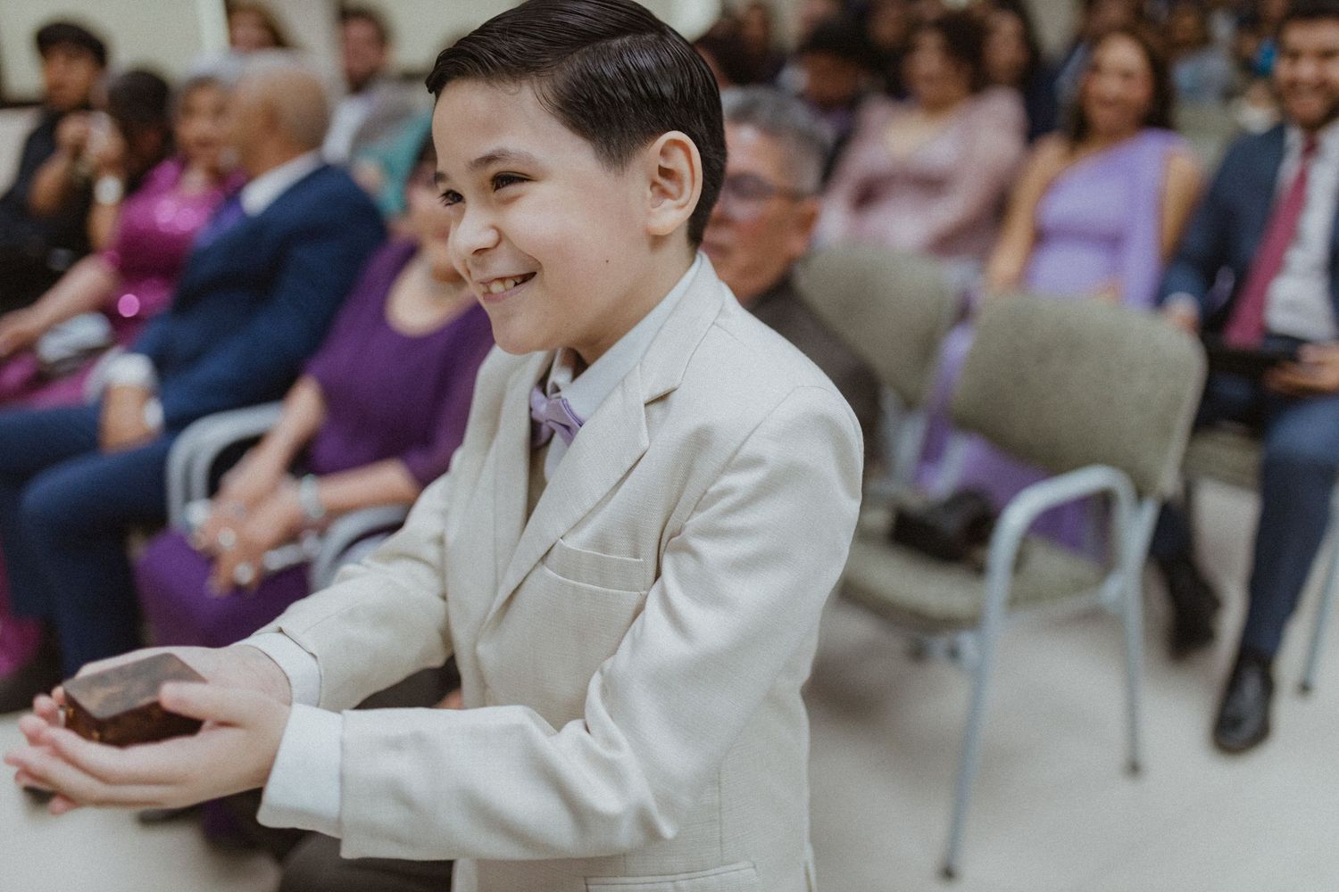 Wedding attendees in formal attire seated in rows during indoor ceremony.