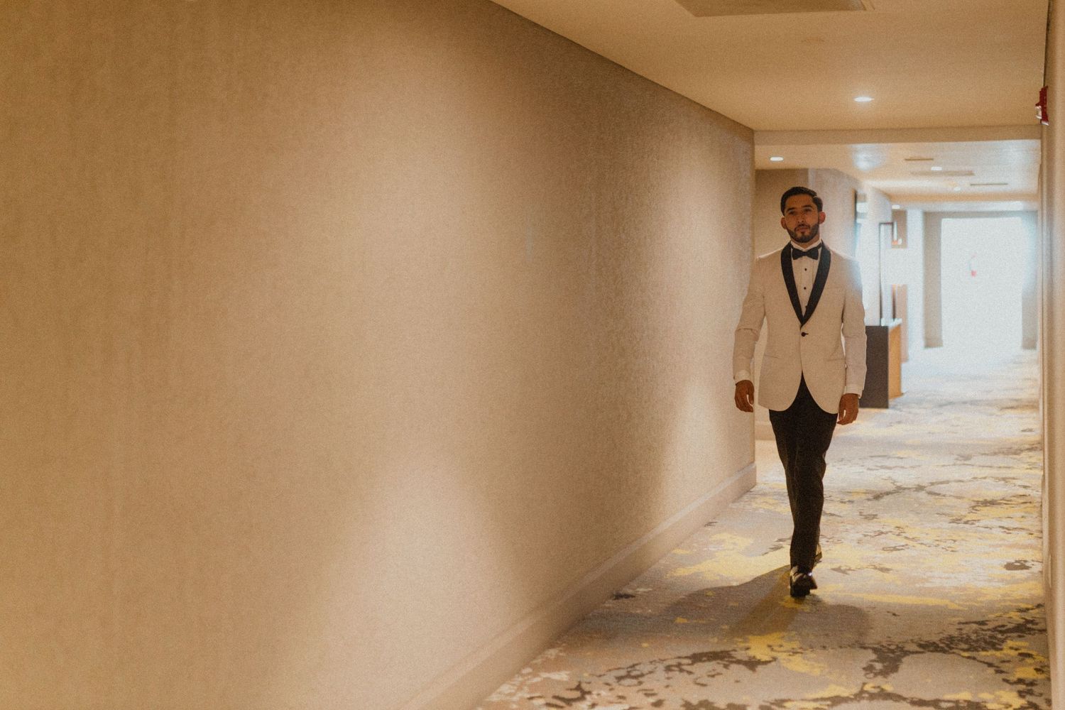 Person in light colored tuxedo walking down elegant hotel hallway.
