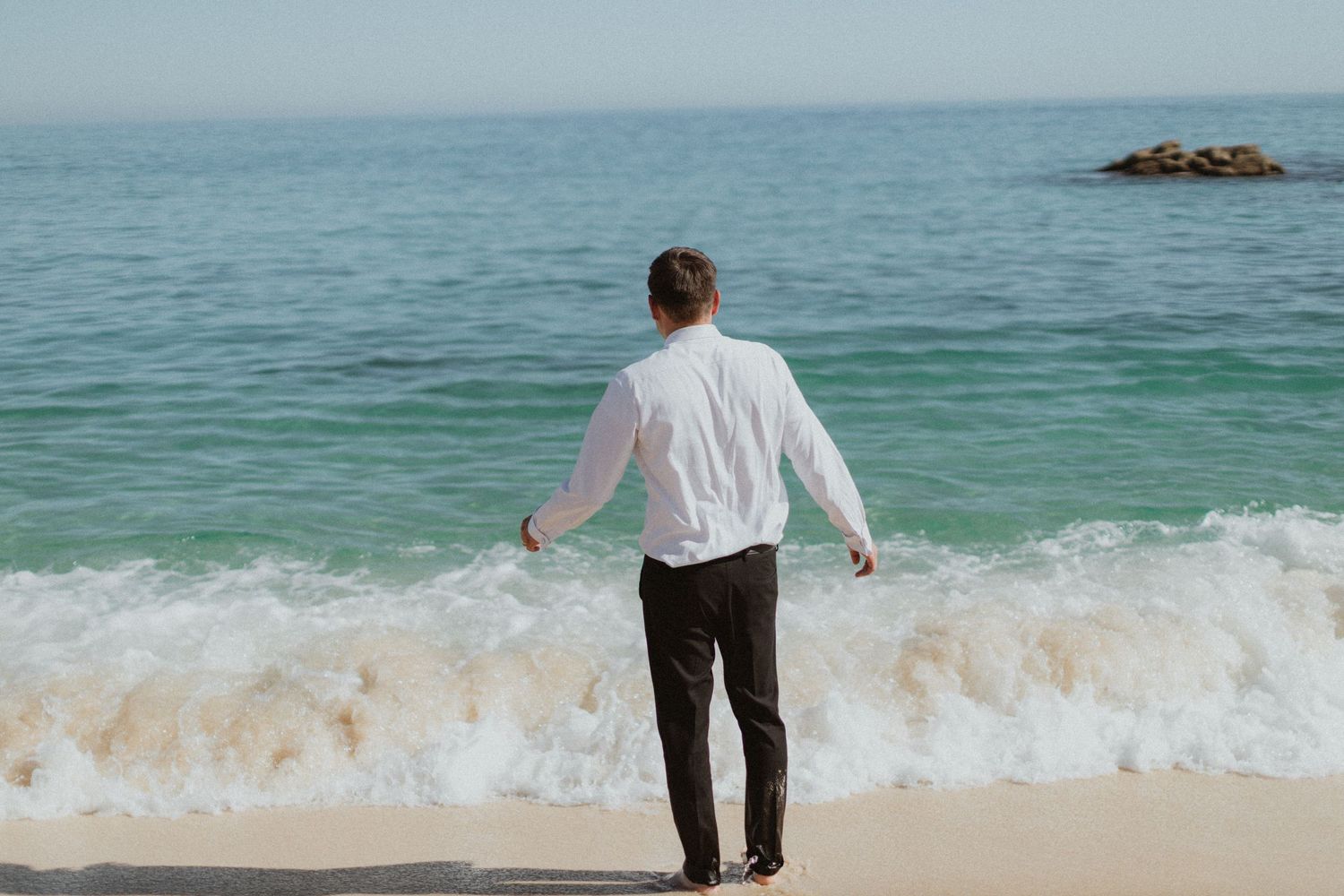 Person in white shirt stands alone facing the ocean waves on a beach.
