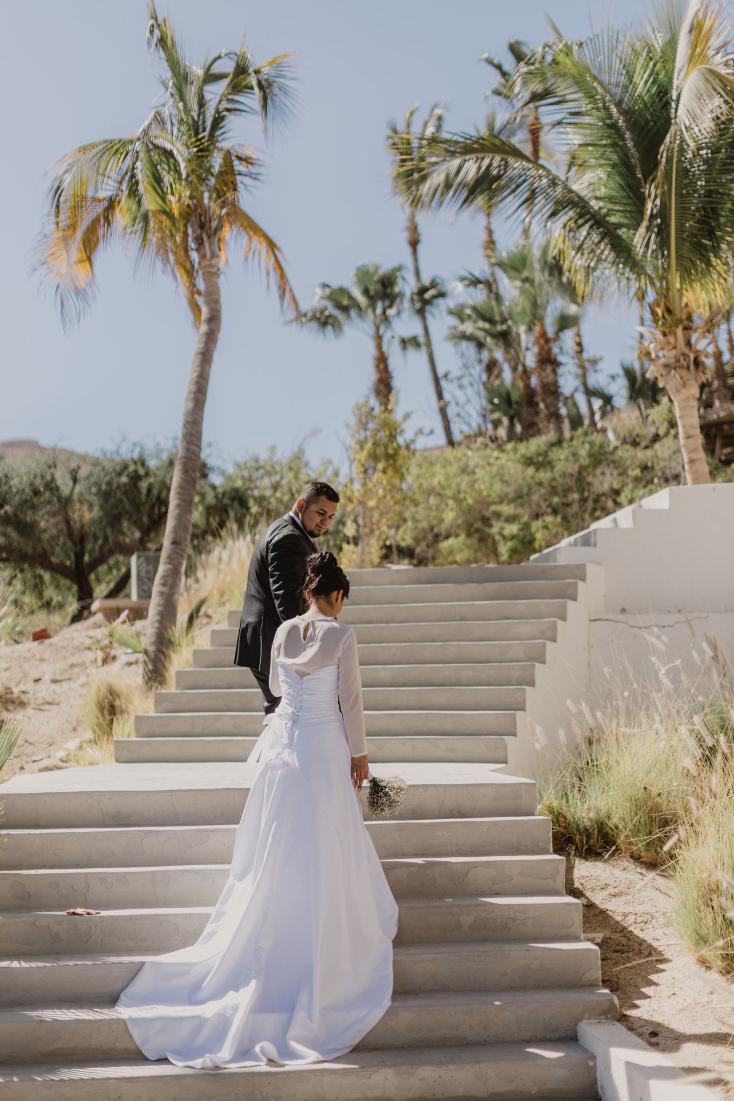 A wedding couple stands on white stairs with palm trees in a tropical destination.