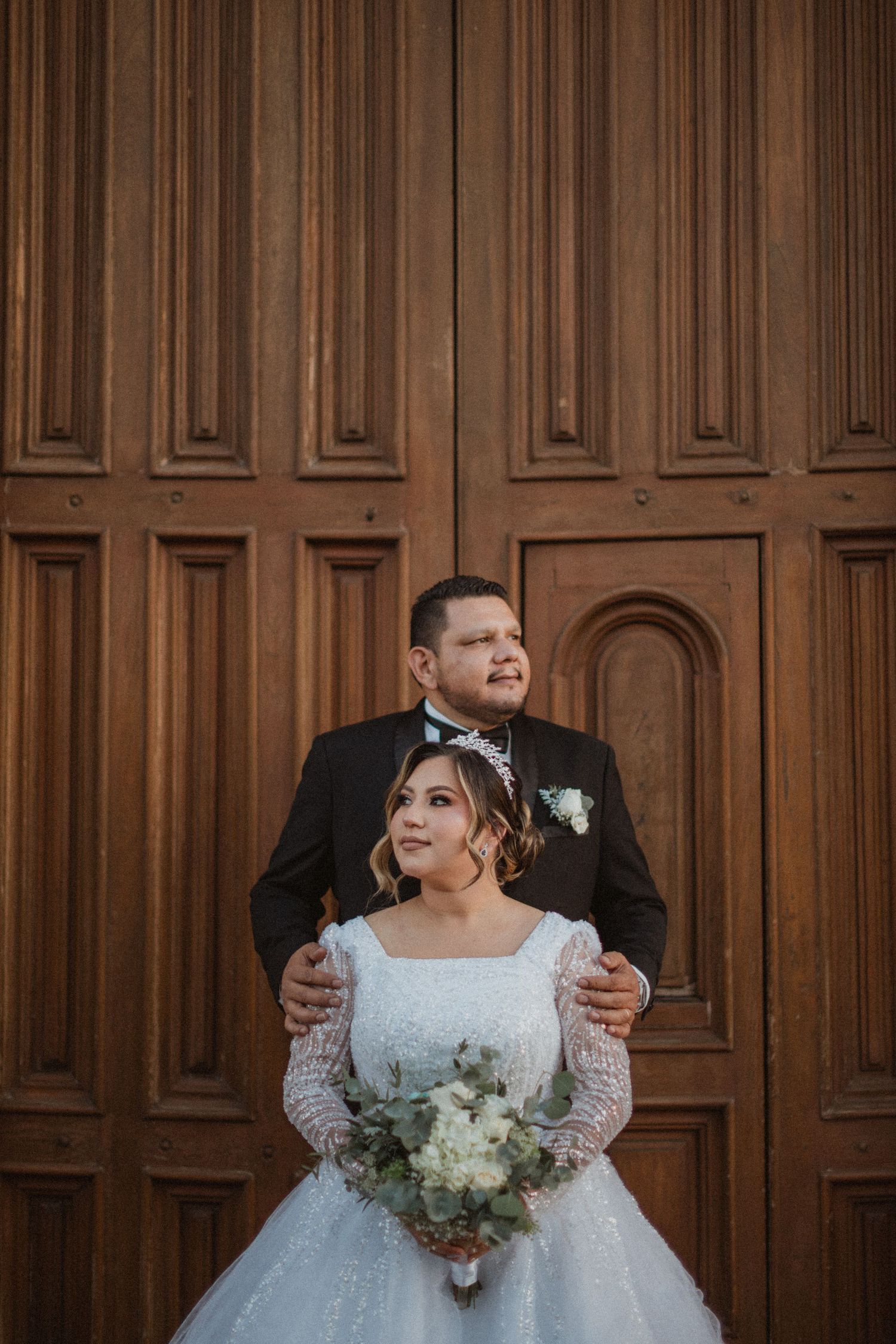 A wedding couple poses against elegant wood paneled walls holding a white floral bouquet.