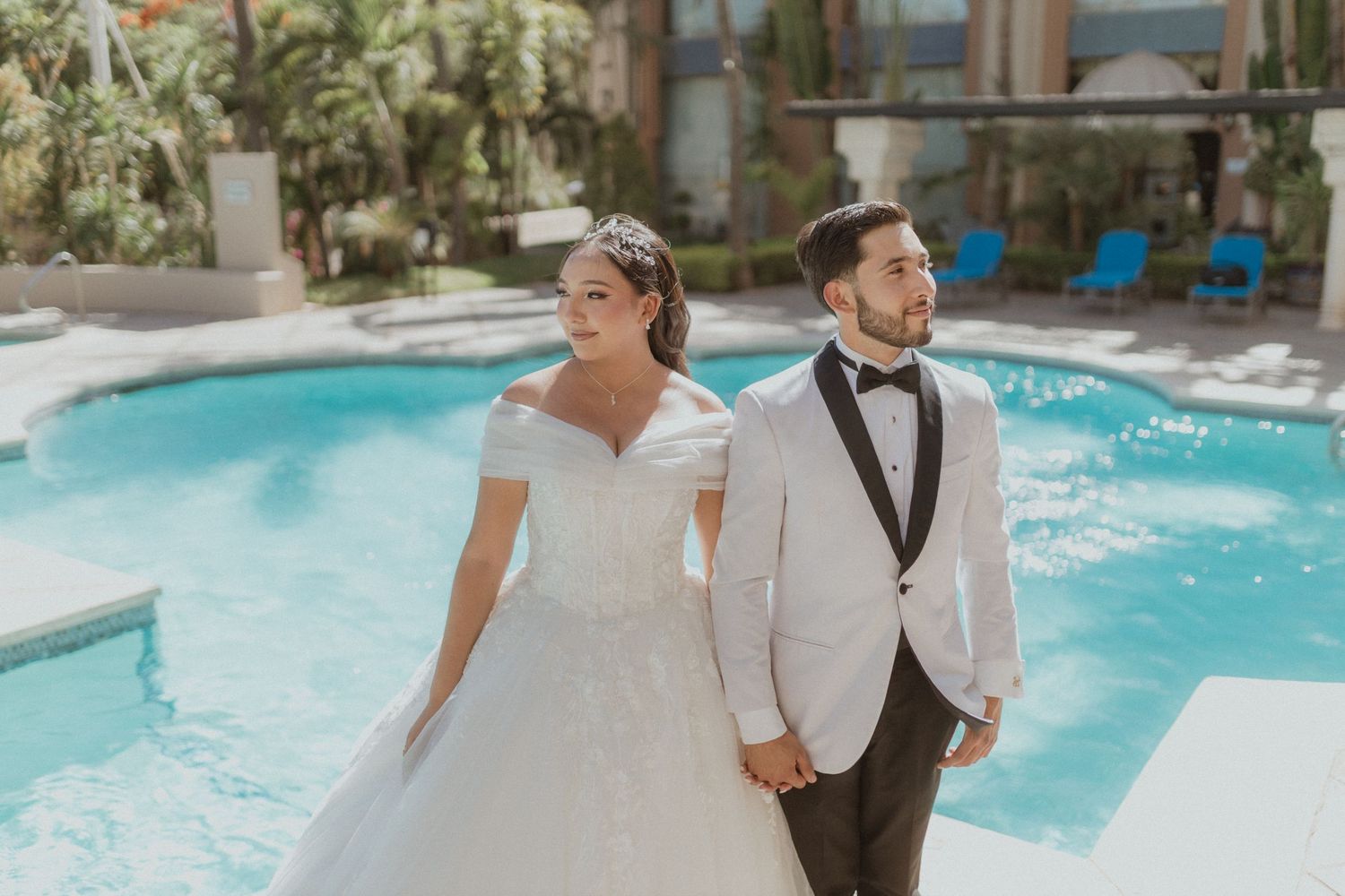 Wedding couple in elegant white attire holding hands by luxury resort pool.