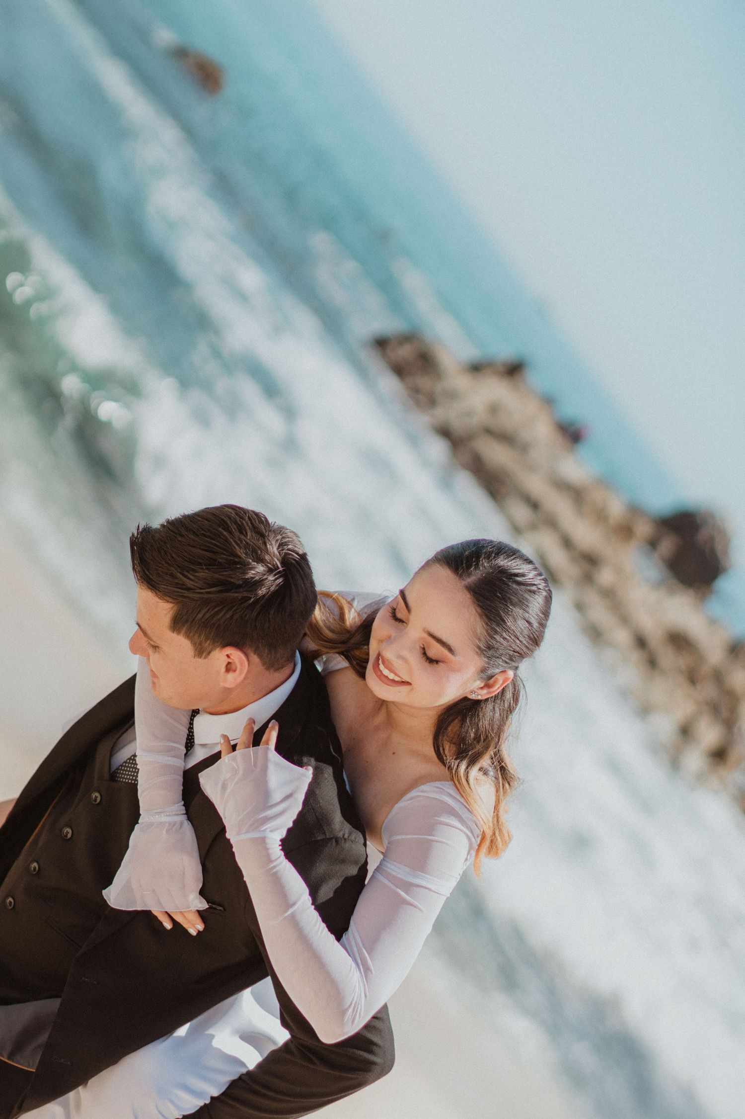 A creative wedding photo angle showing a couple dancing on the beach with crashing waves.