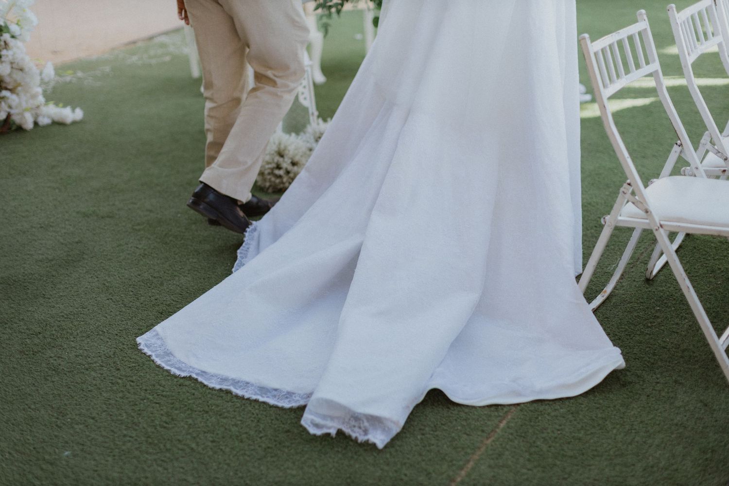 Close-up of a flowing white wedding dress train on green grass with white chairs in the background.