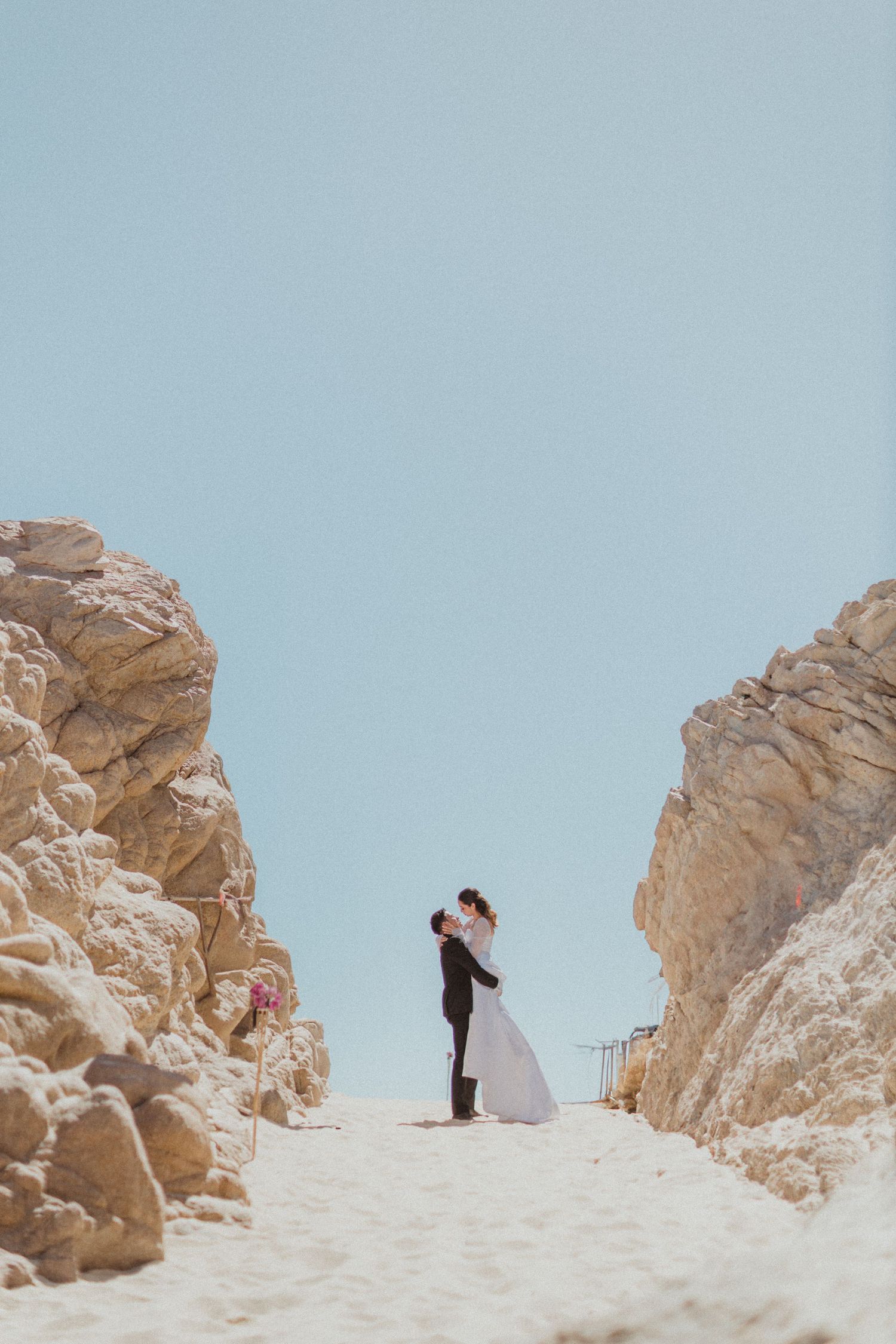 A romantic wedding couple embraces between towering rocky cliffs against a bright blue sky.
