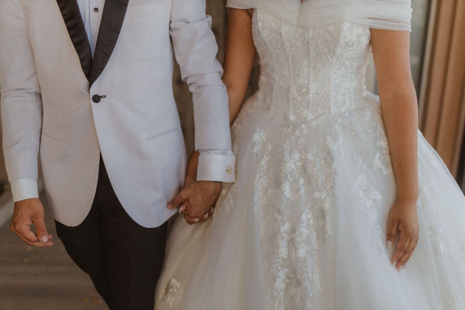 Close-up of couple in wedding attire holding hands, showing white tuxedo jacket and lace gown details.