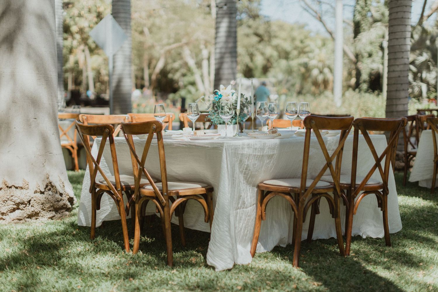 Elegant outdoor wedding reception setup with wooden chairs and white decor under palm trees.