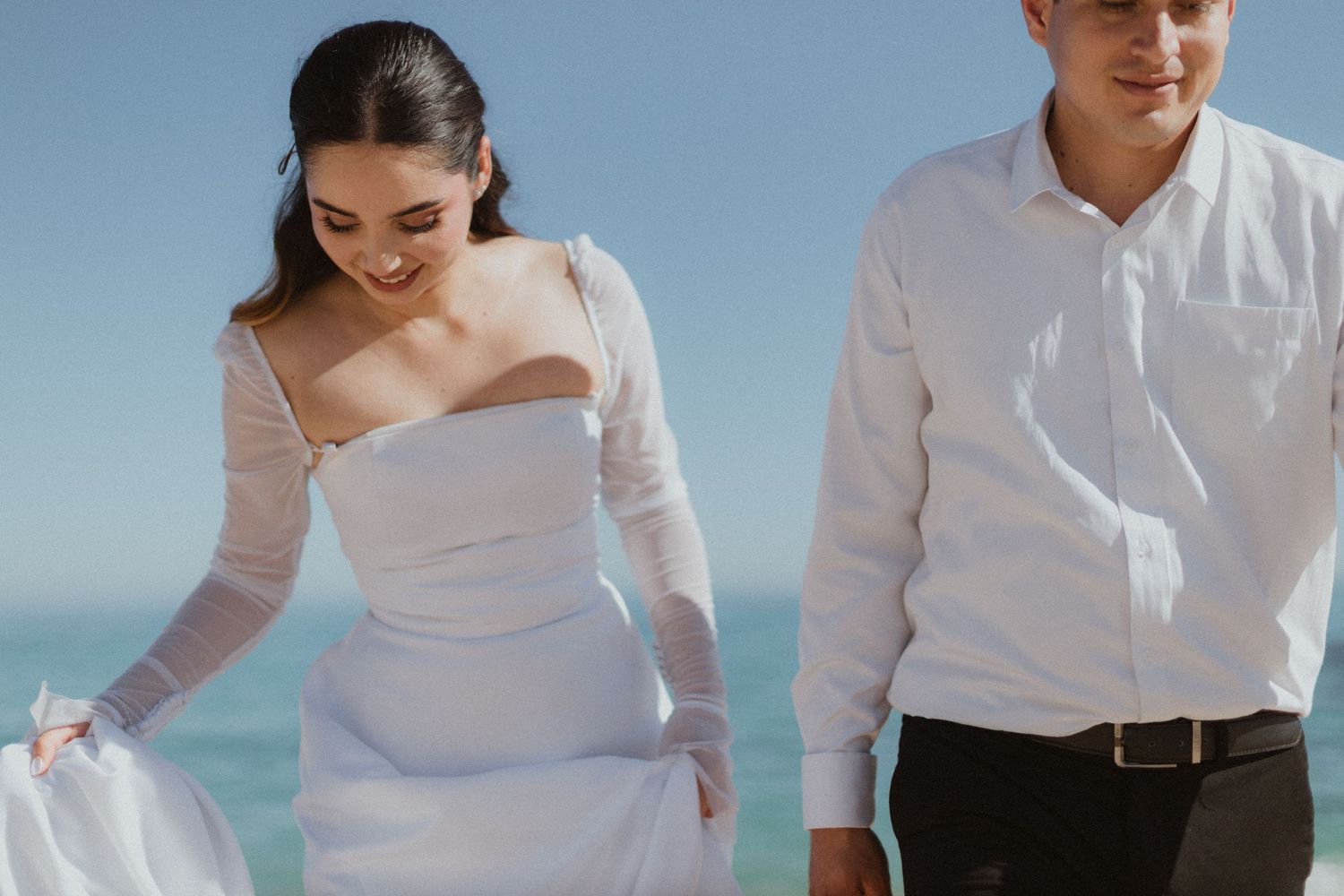 A stunning beachside portrait of a bride in a white dress and groom in white shirt against ocean backdrop.