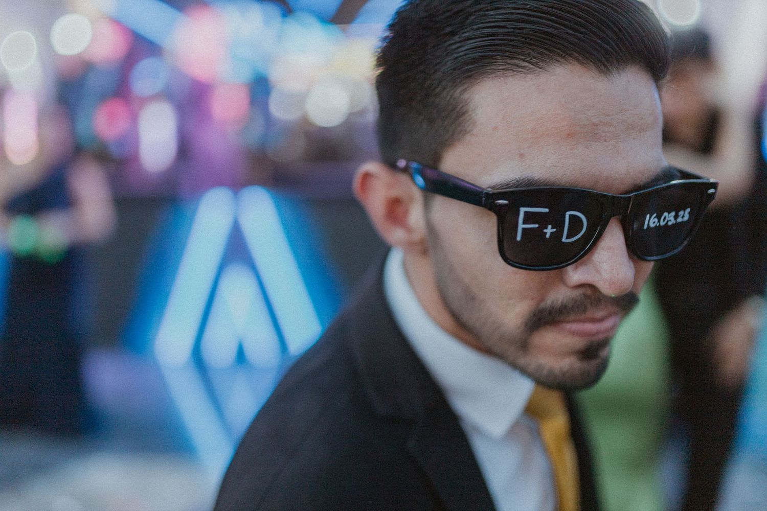 Professional party DJ wearing dark aviator sunglasses plays music at an event with colorful lights in background.