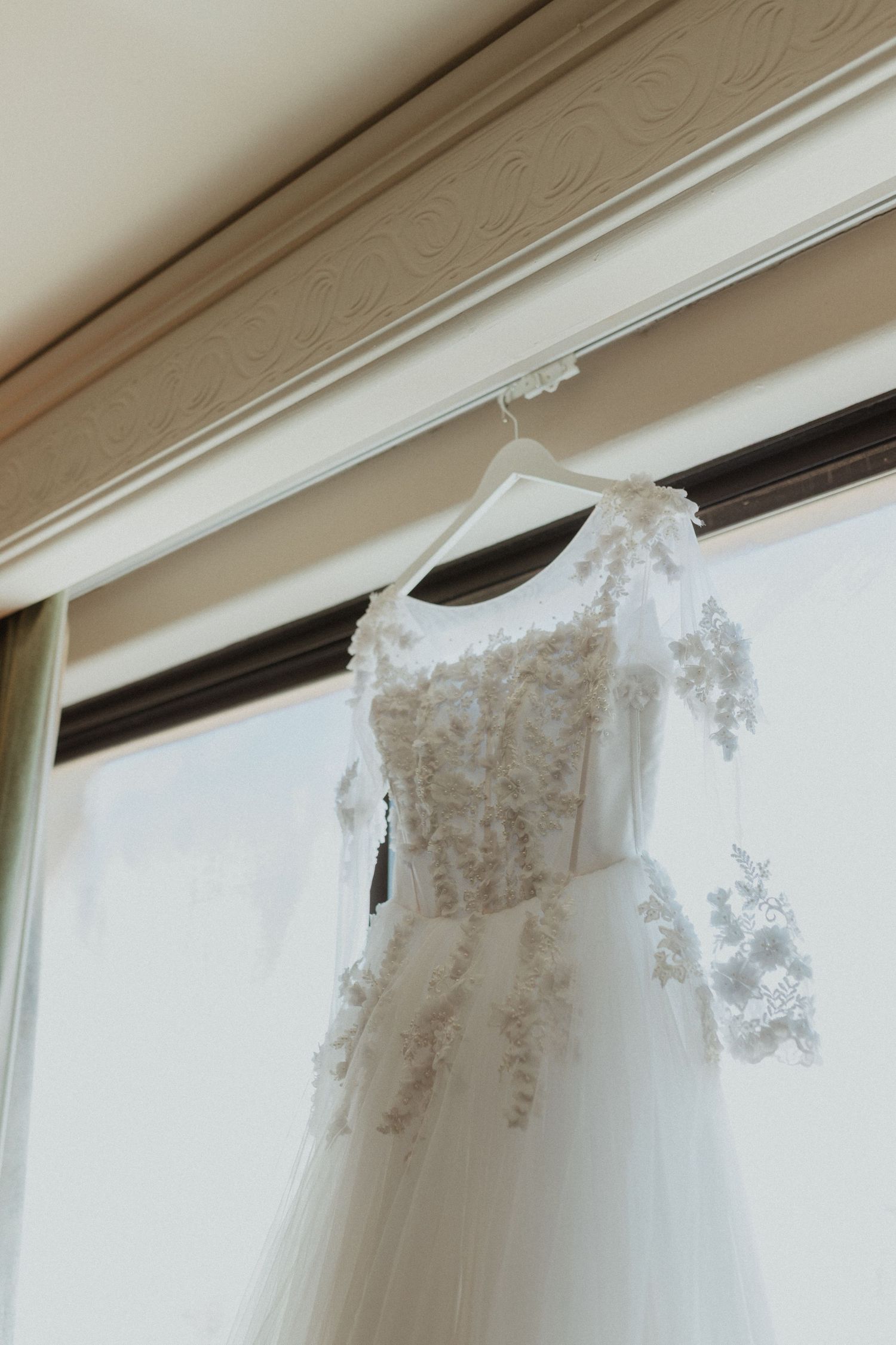 Elegant white lace wedding dress hanging by window with ornate molding.