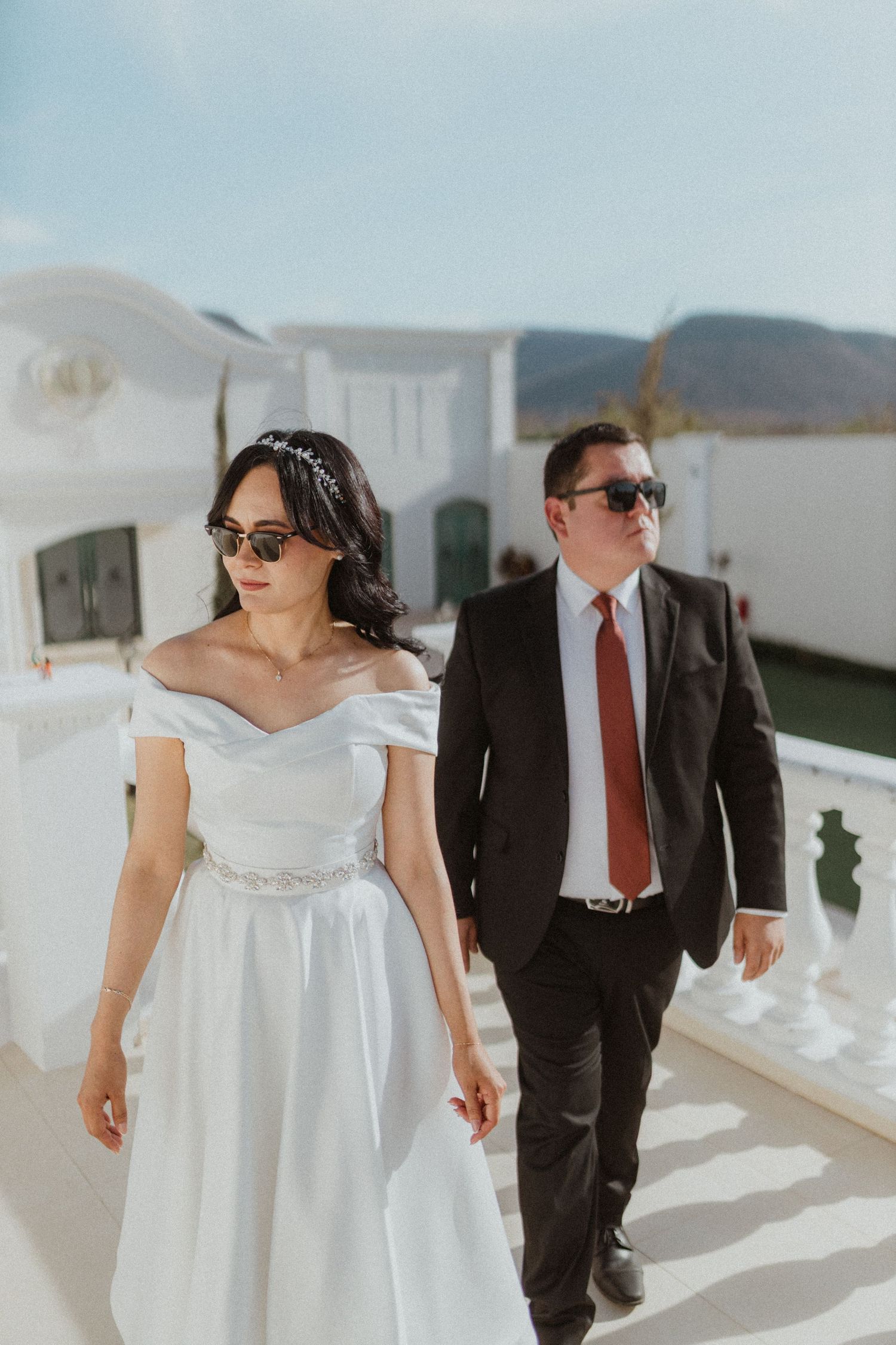 Wedding couple in sunglasses walking together on white terrace against blue sky.