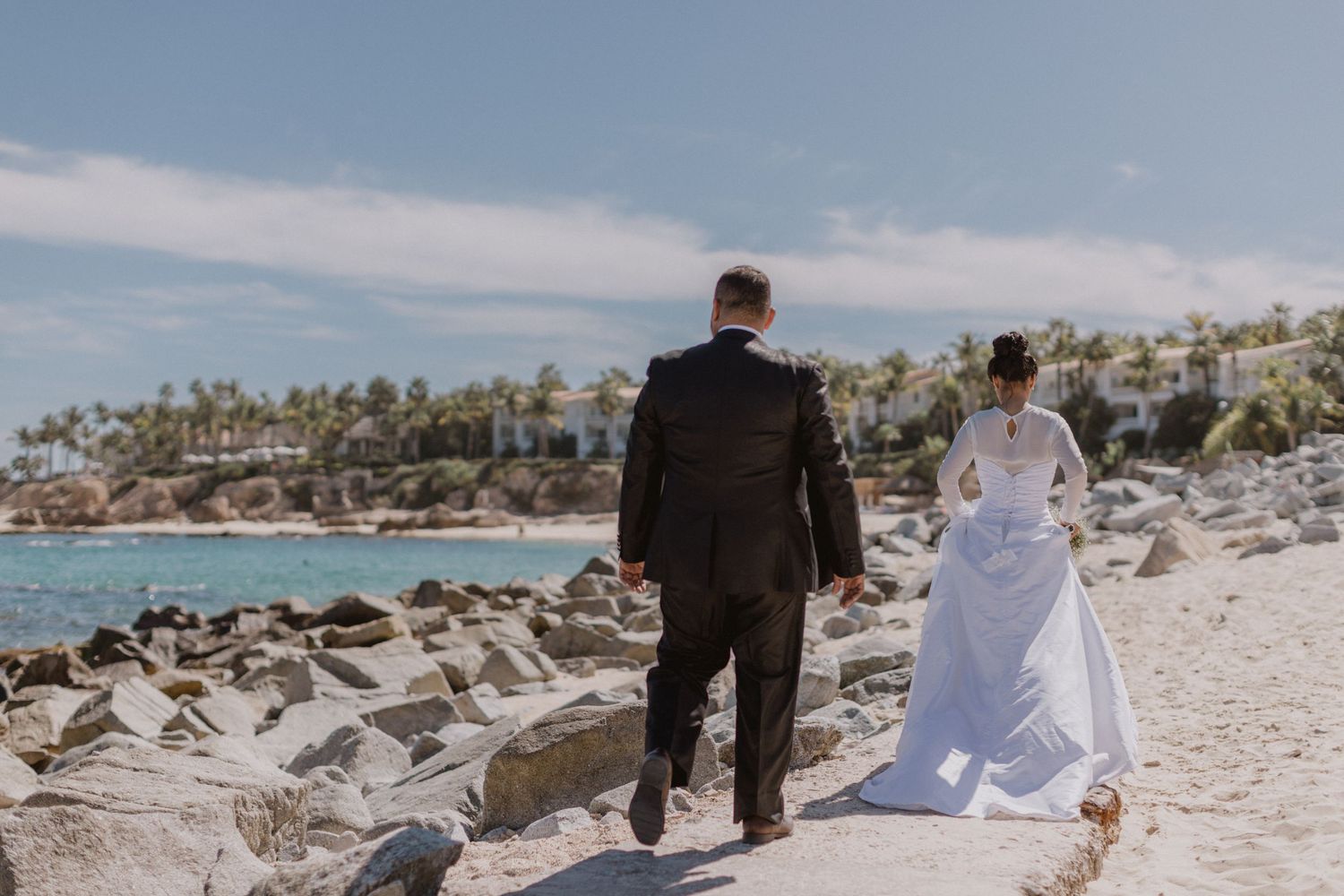 Newlyweds walking together on rocky beach with palm trees in background.
