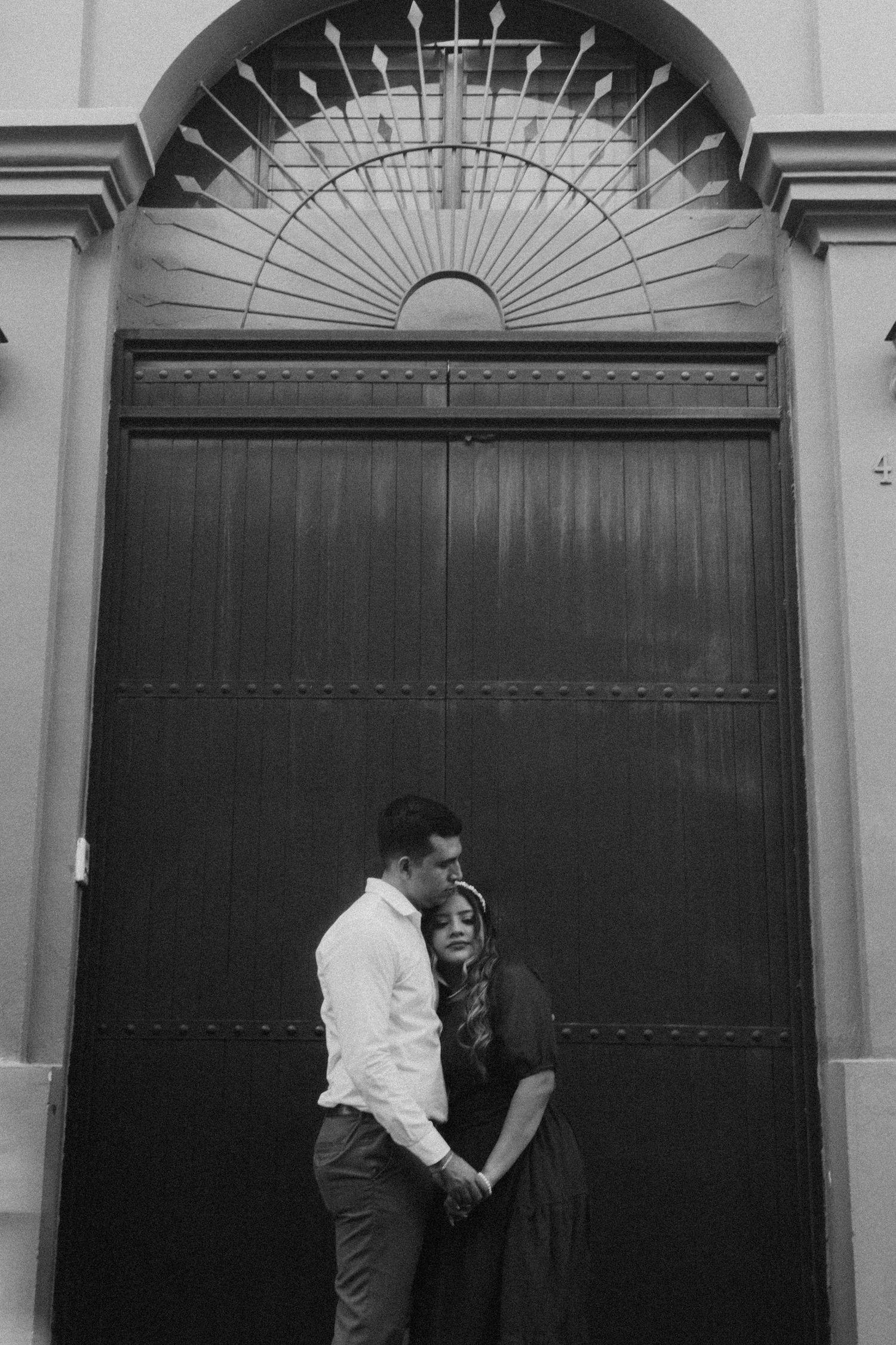 A romantic couple poses against a large wooden door with an ornate fan window above.