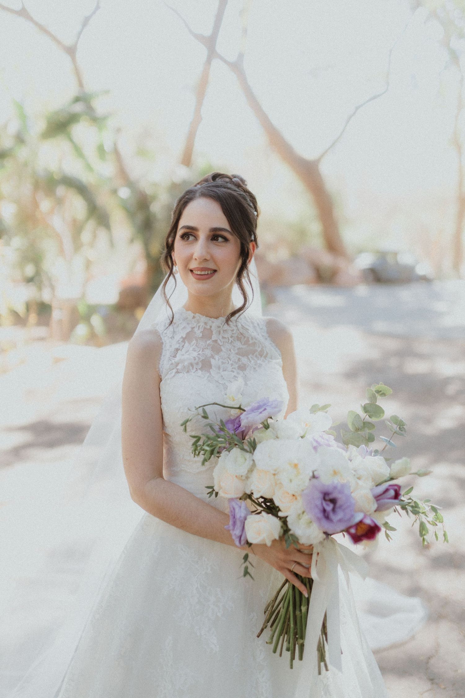 Bride in lace dress holds elegant purple and white floral bouquet outdoors.