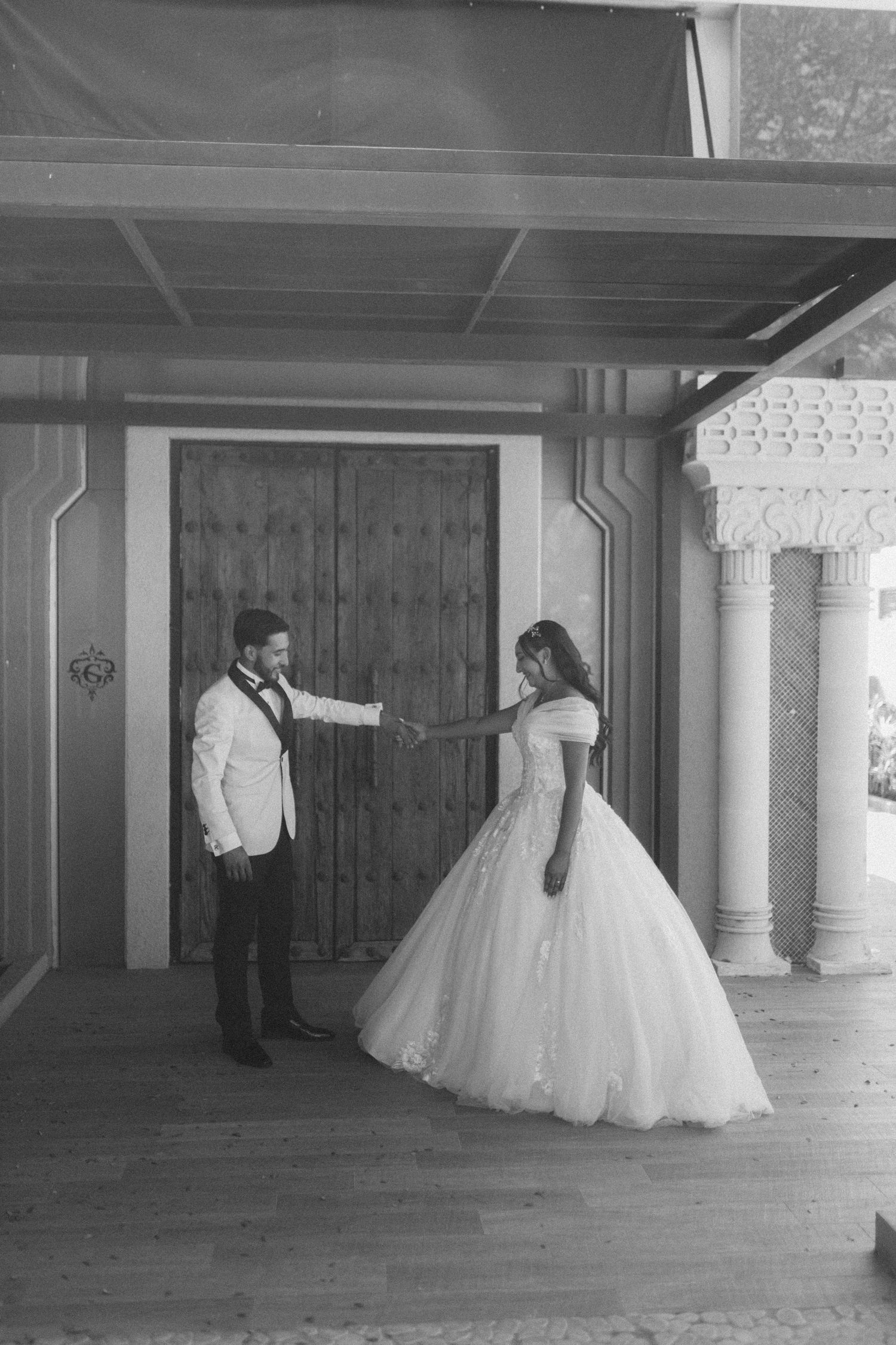 Black and white wedding photo of a couple in formal attire posing in front of a rustic wooden door.