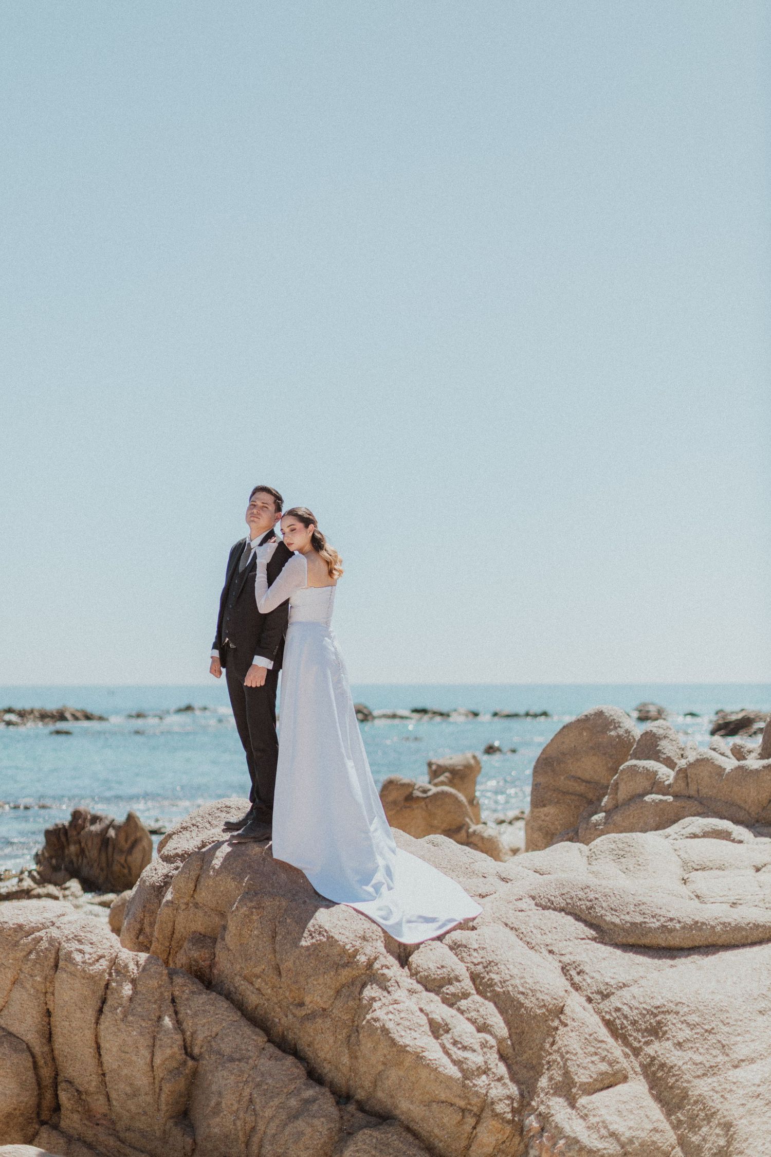 Wedding couple poses on rocky coastal outcrop against blue sky.