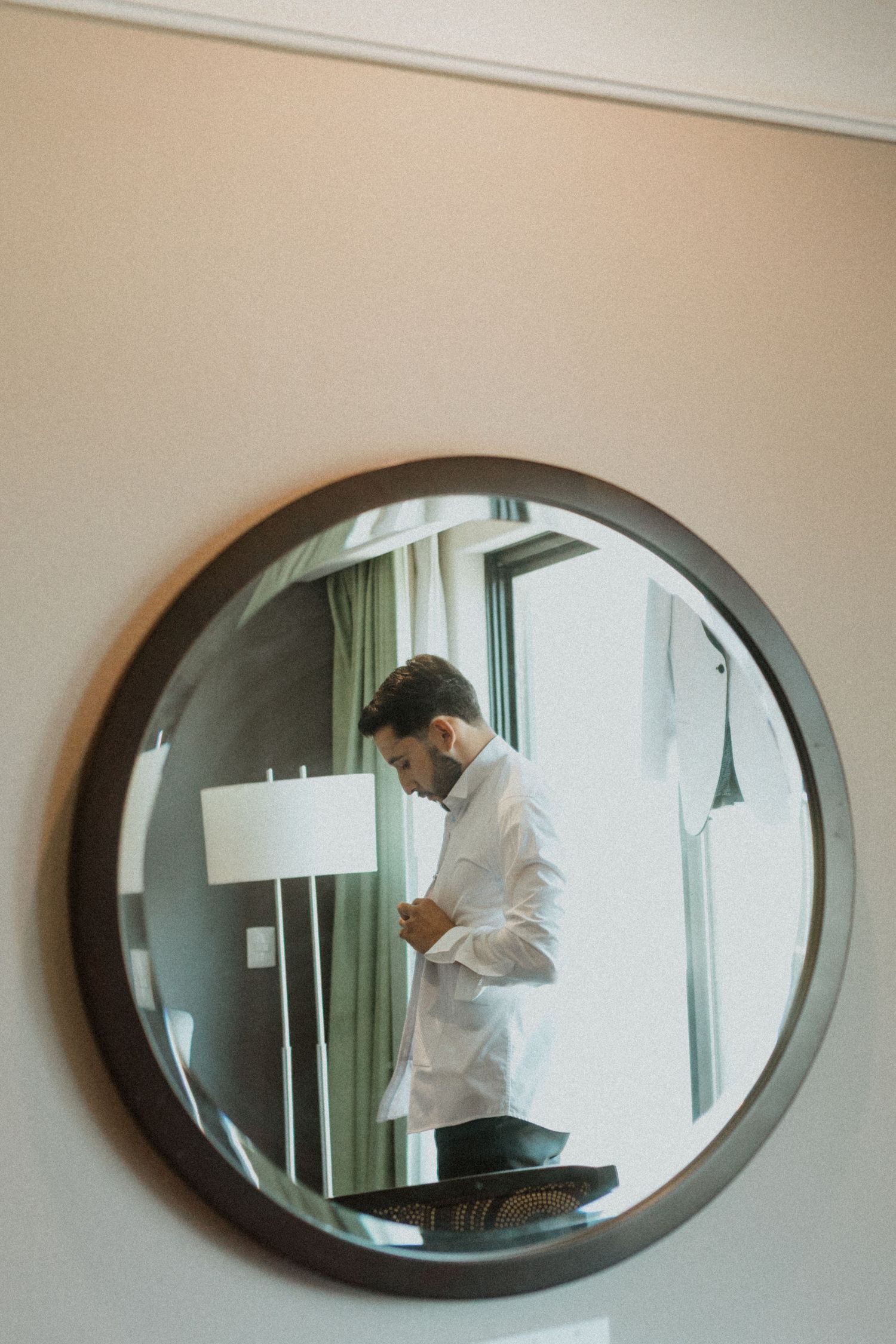 Reflection in a round mirror shows a person adjusting formal attire near a window.
