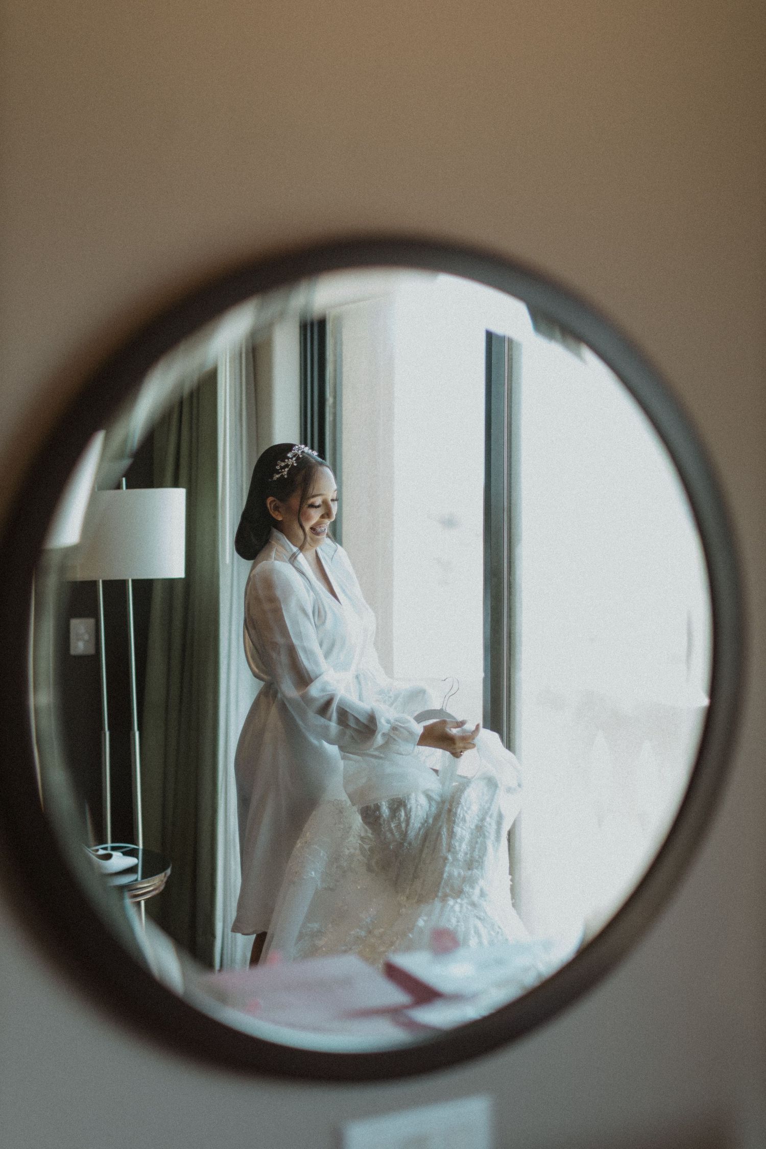 A person in a white robe looks at a wedding dress through an oval mirror reflection.