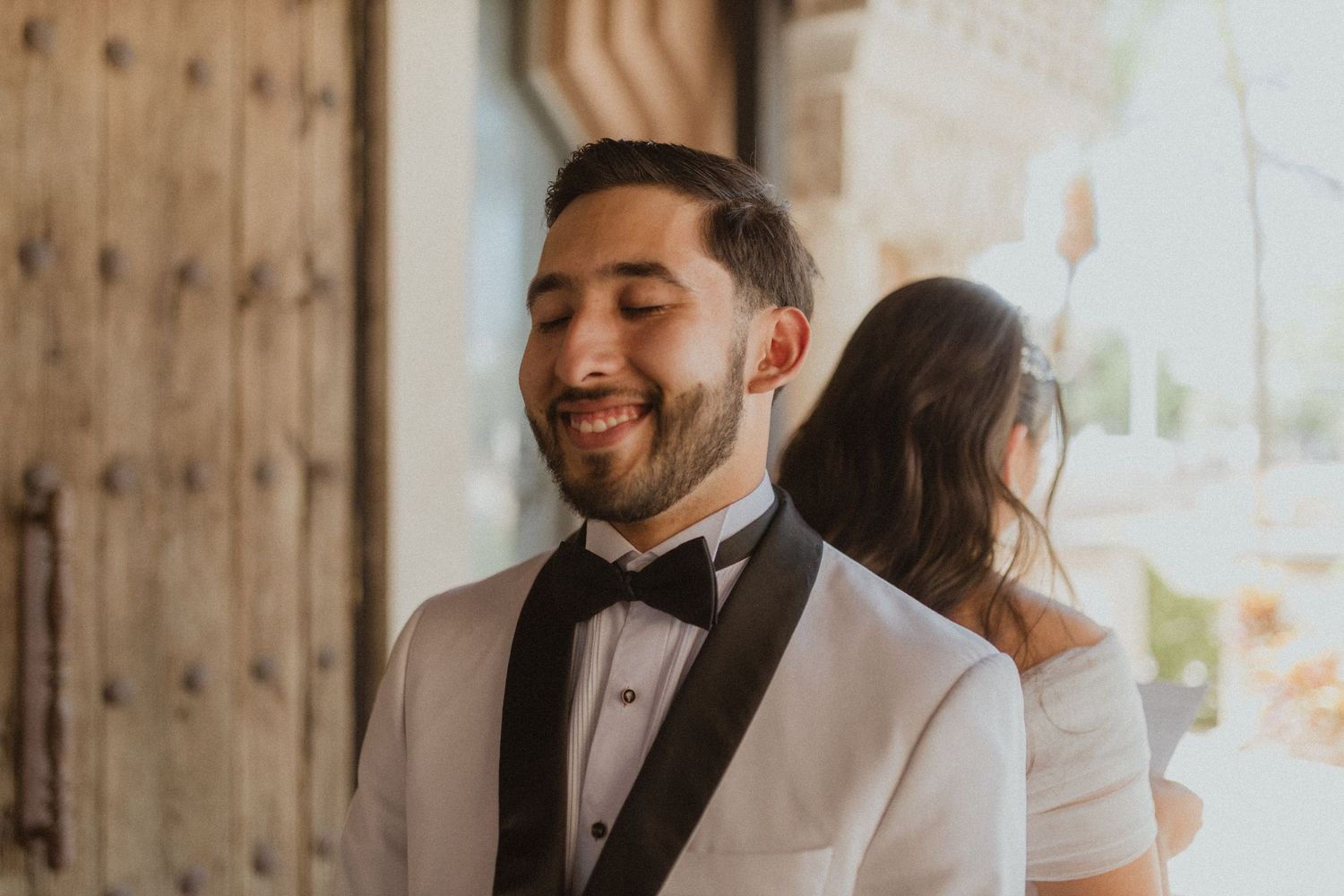 A groom in a white tuxedo jacket and black bow tie smiles during his wedding first look.