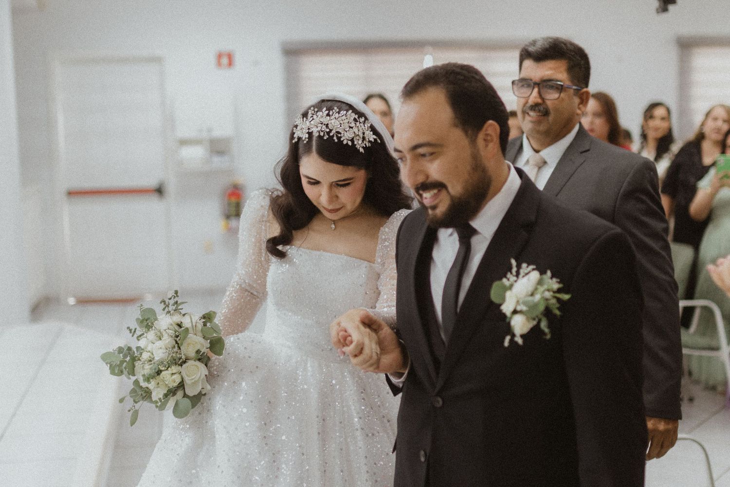 Bride in a sparkly white gown and groom in black suit walk together joyfully down a corridor.