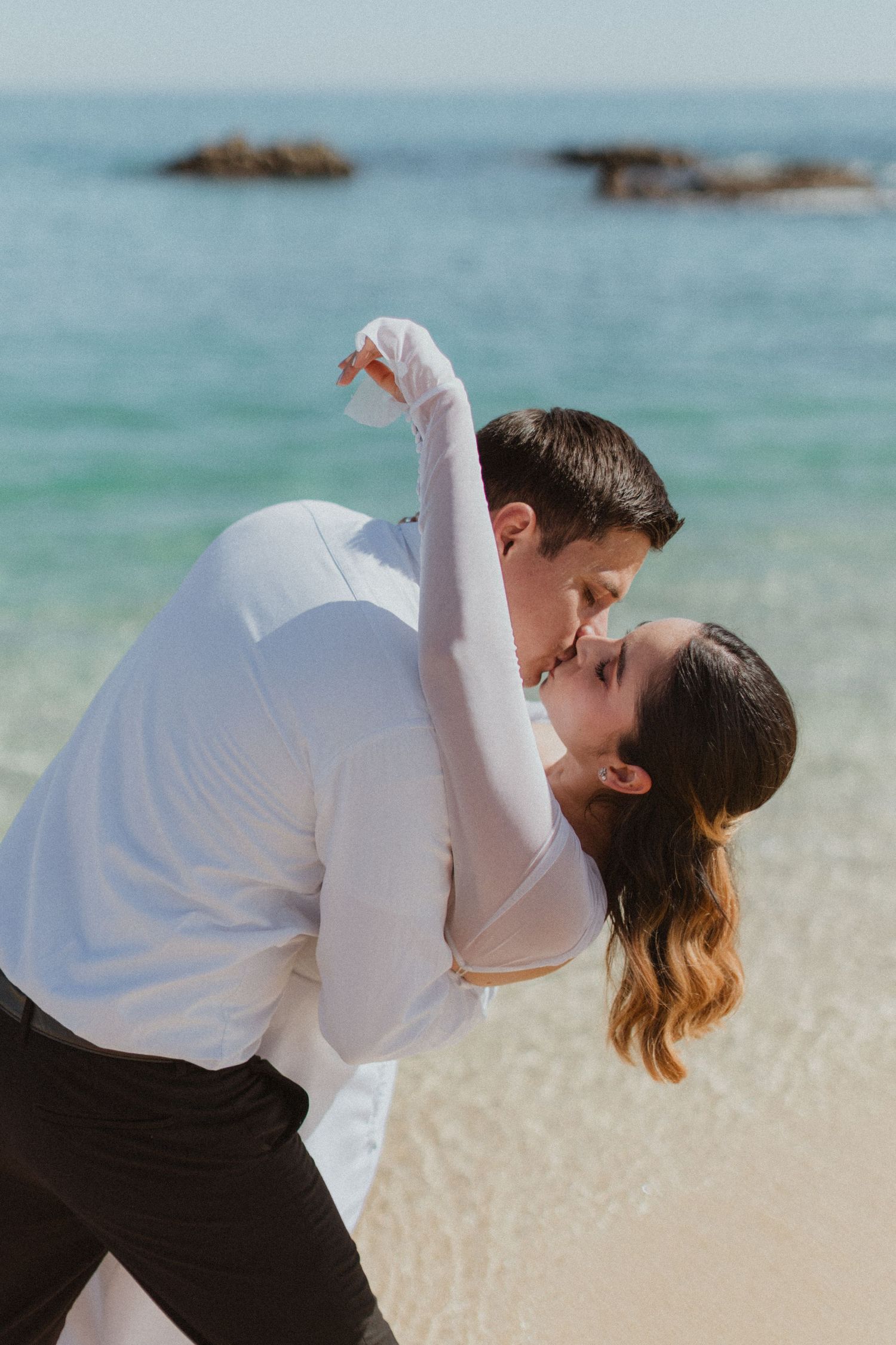 A romantic dip pose on the beach with waves and rocks in the background.