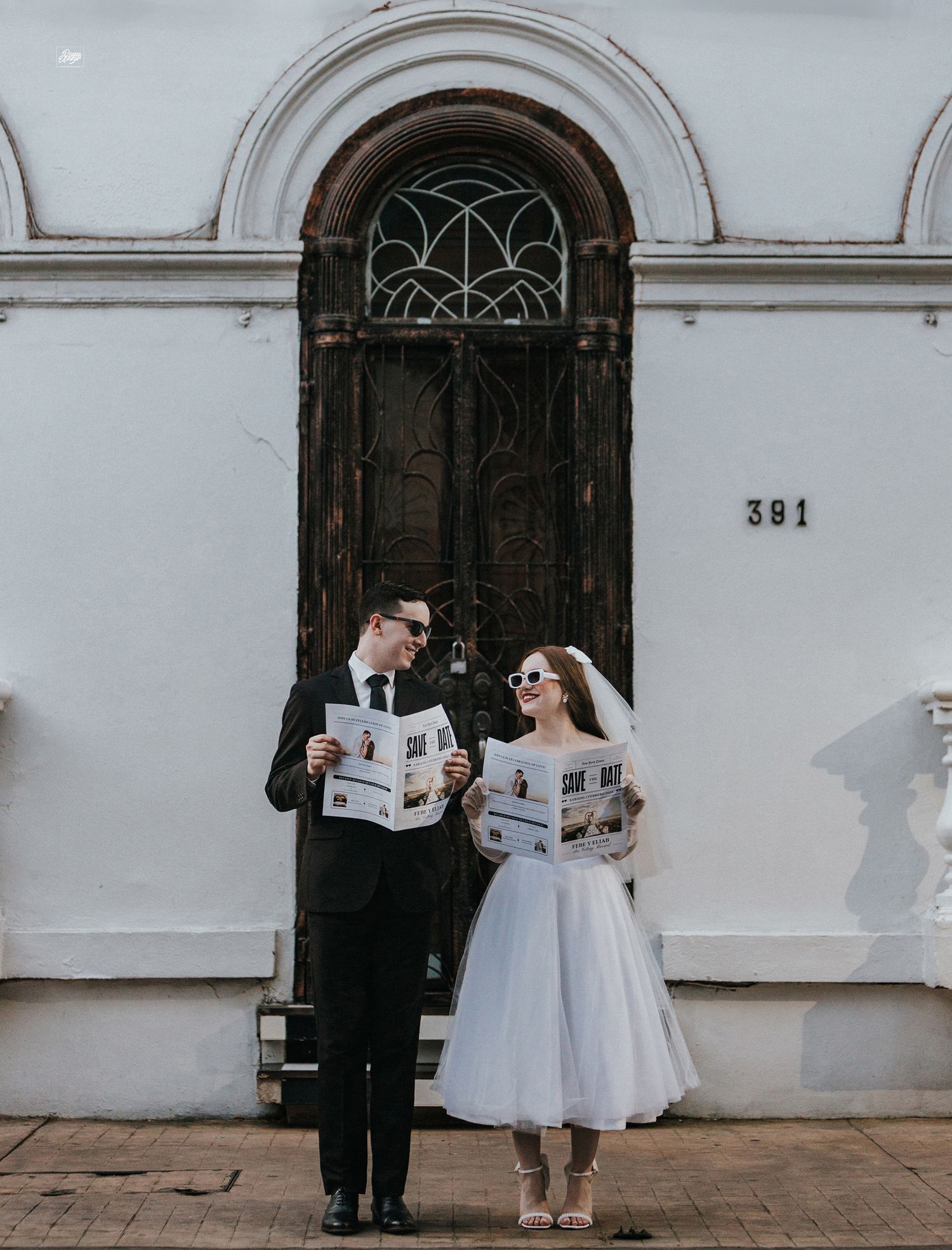 A newlywed couple reads newspapers together in front of an ornate vintage doorway.