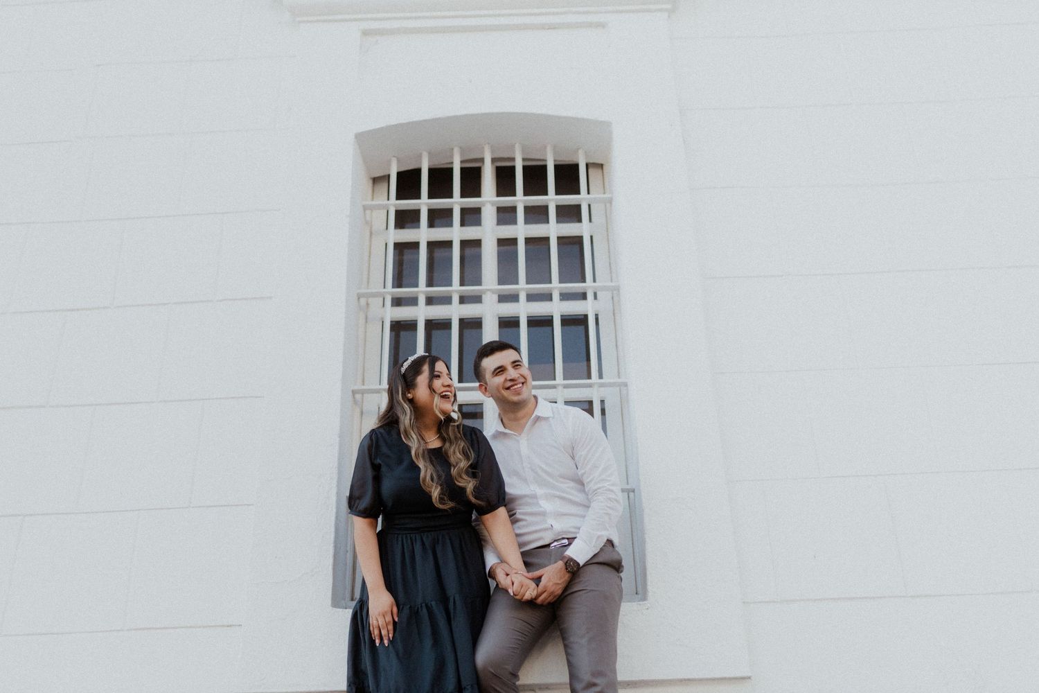 A couple shares a playful moment against a white building with barred windows.