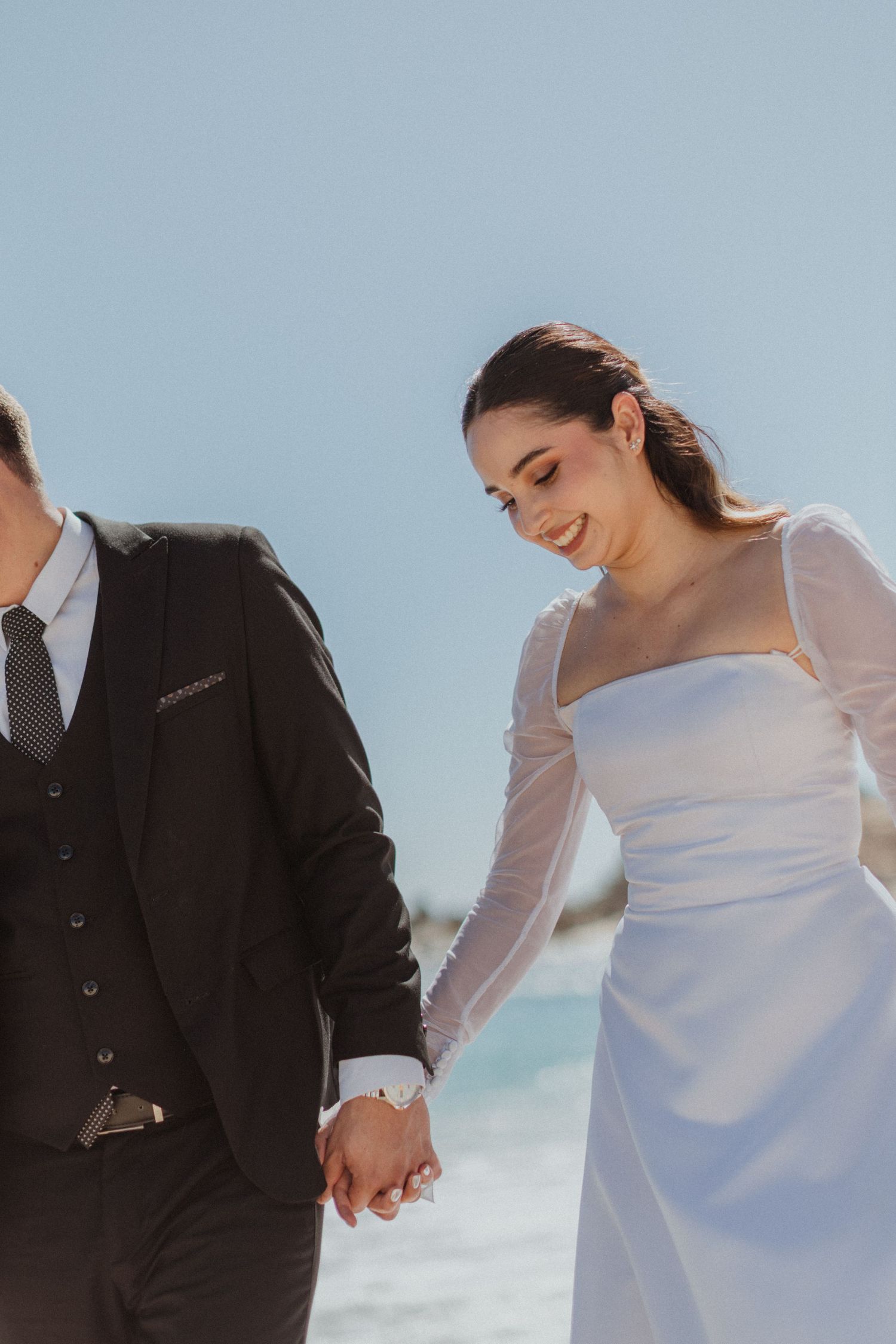 A couple in wedding attire holds hands while walking along a sunny beach.