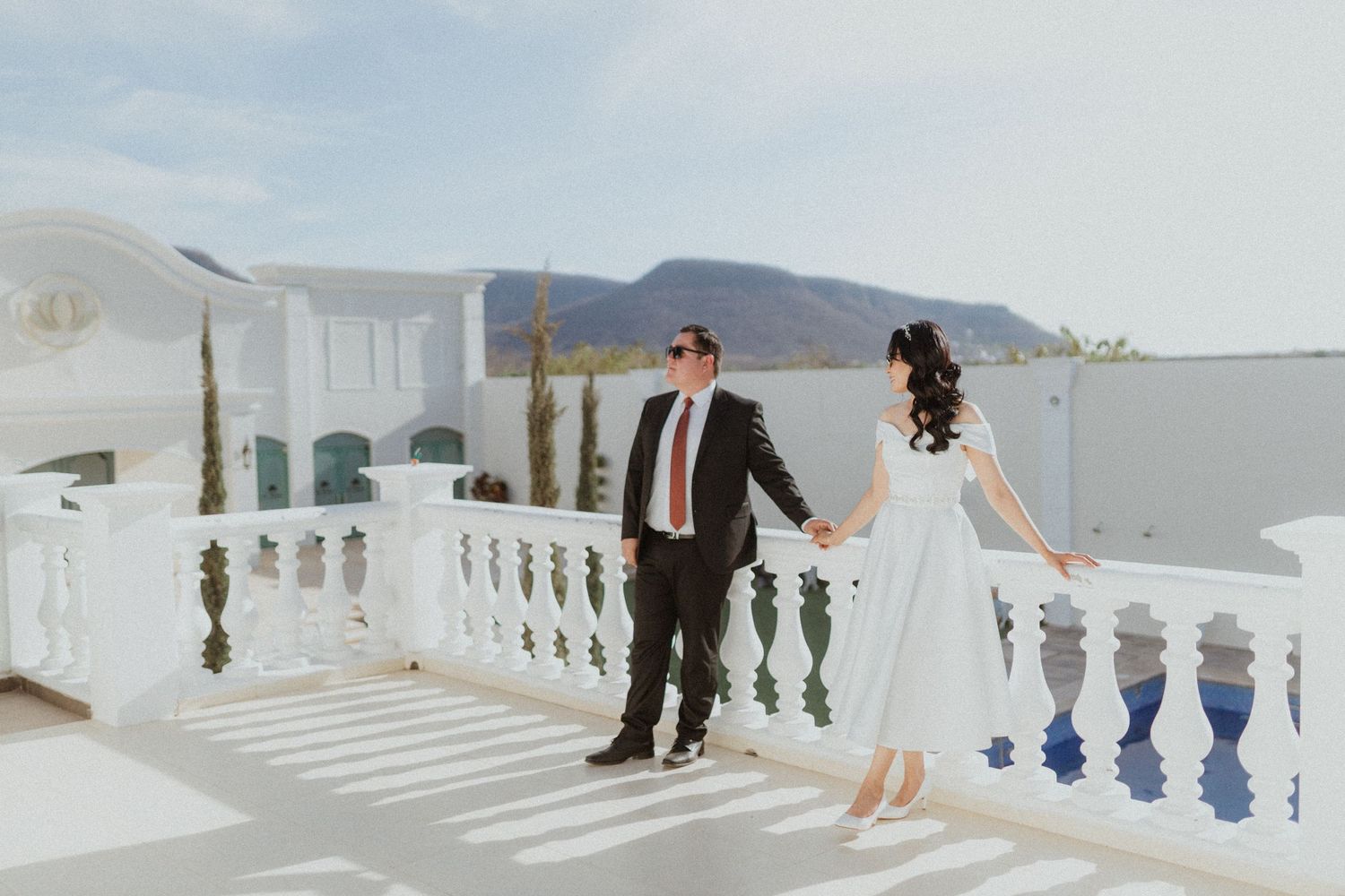 Couple in wedding attire posing on a white balcony overlooking mountains in Santorini.