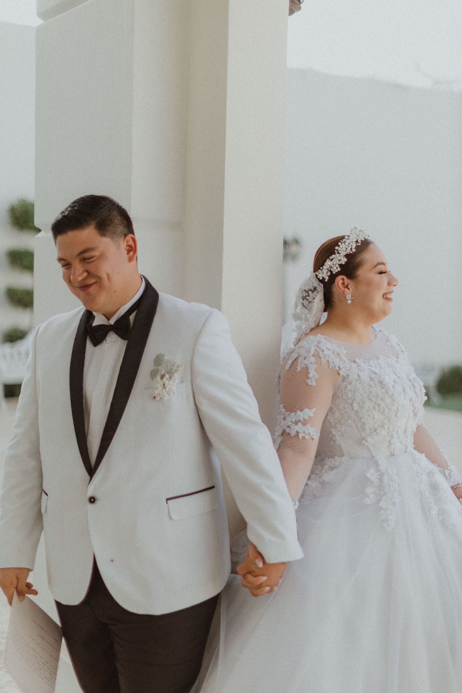 A newlywed couple holding hands and laughing in a white hallway during their wedding.